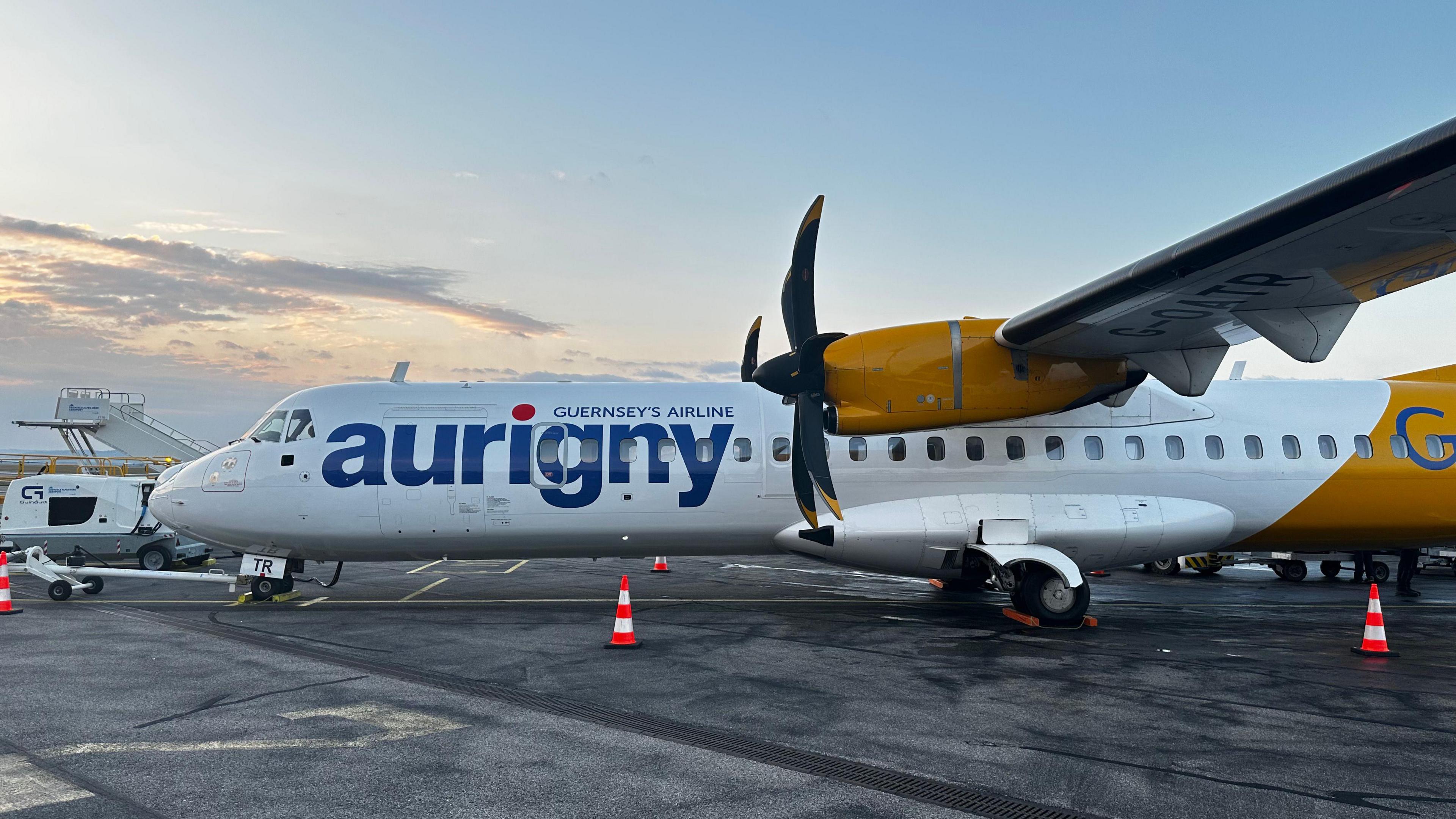 A white and yellow aurigny ATR aircraft on the tarmac in Guernsey. There are cones around the plane.