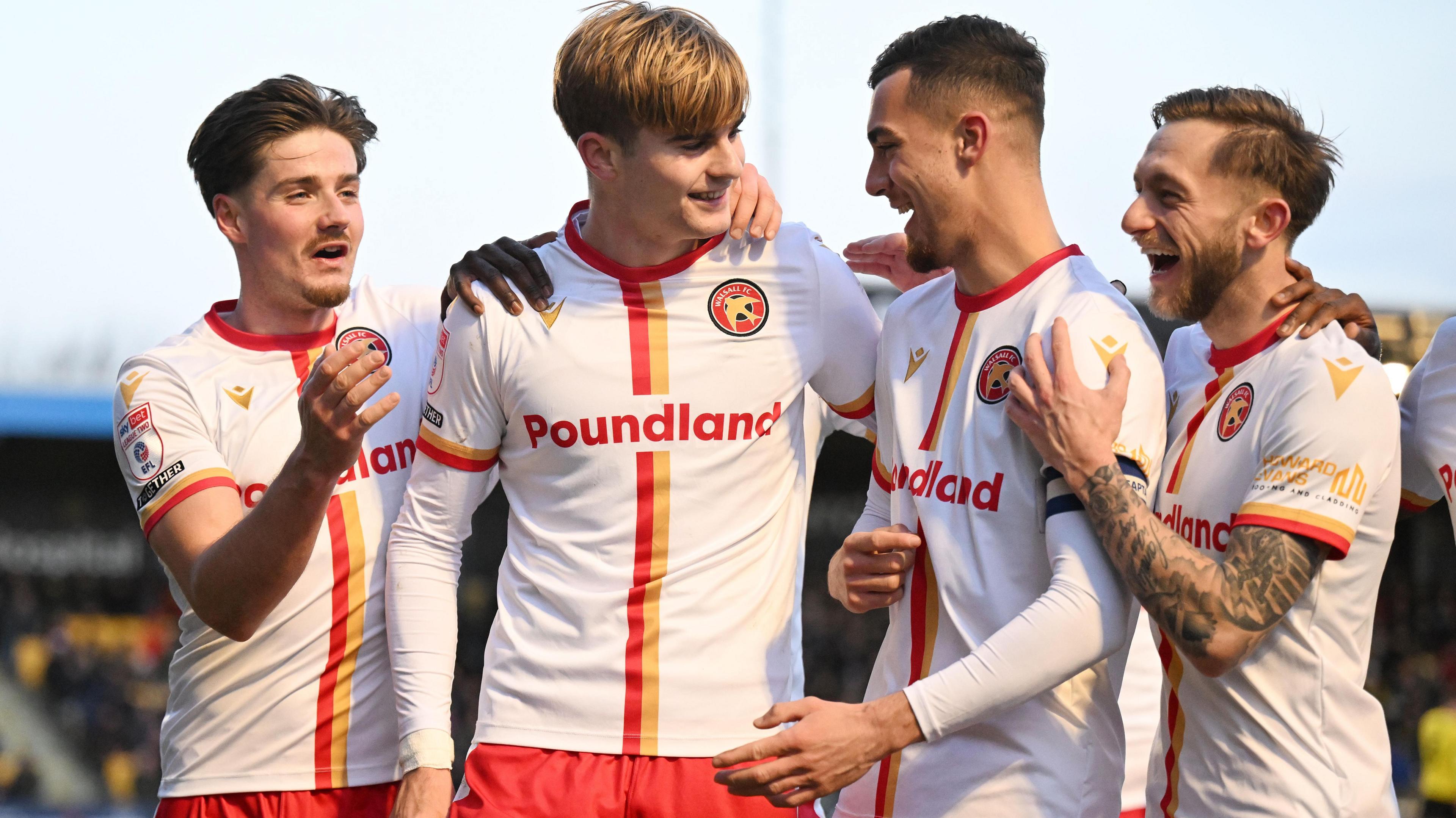 Nathan Lowe, wearing a white kit with red and yellow trim and sporting a floppy light brown hairstyle, celebrates a goal with three Walsall team-mates