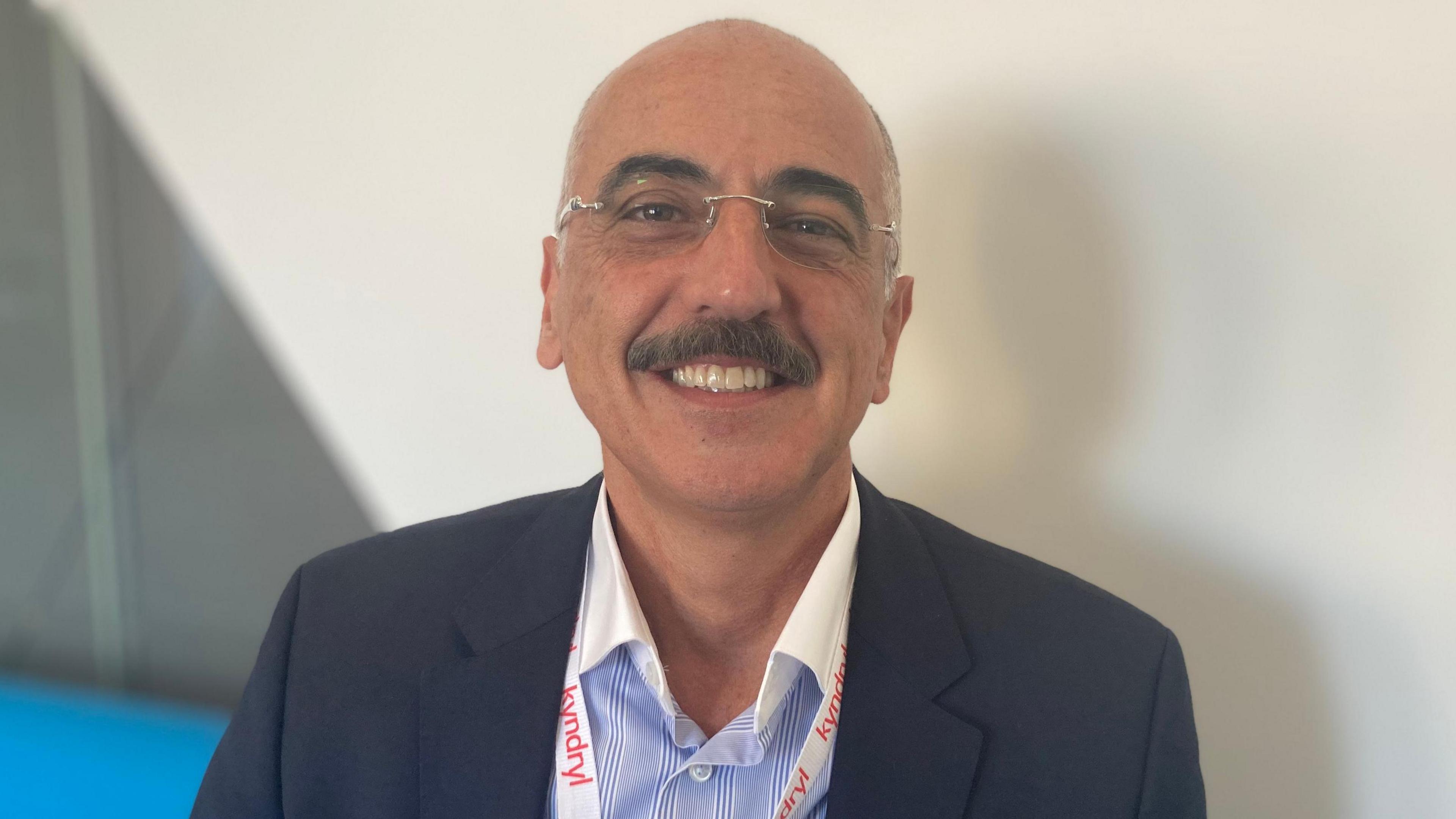 Antoine Shagoury is smiling at the camera. He has short hair and a moustache and is wearing glasses and a blue and white shirt, black suit jacket and a white lanyard with Kyndryl written in red text.