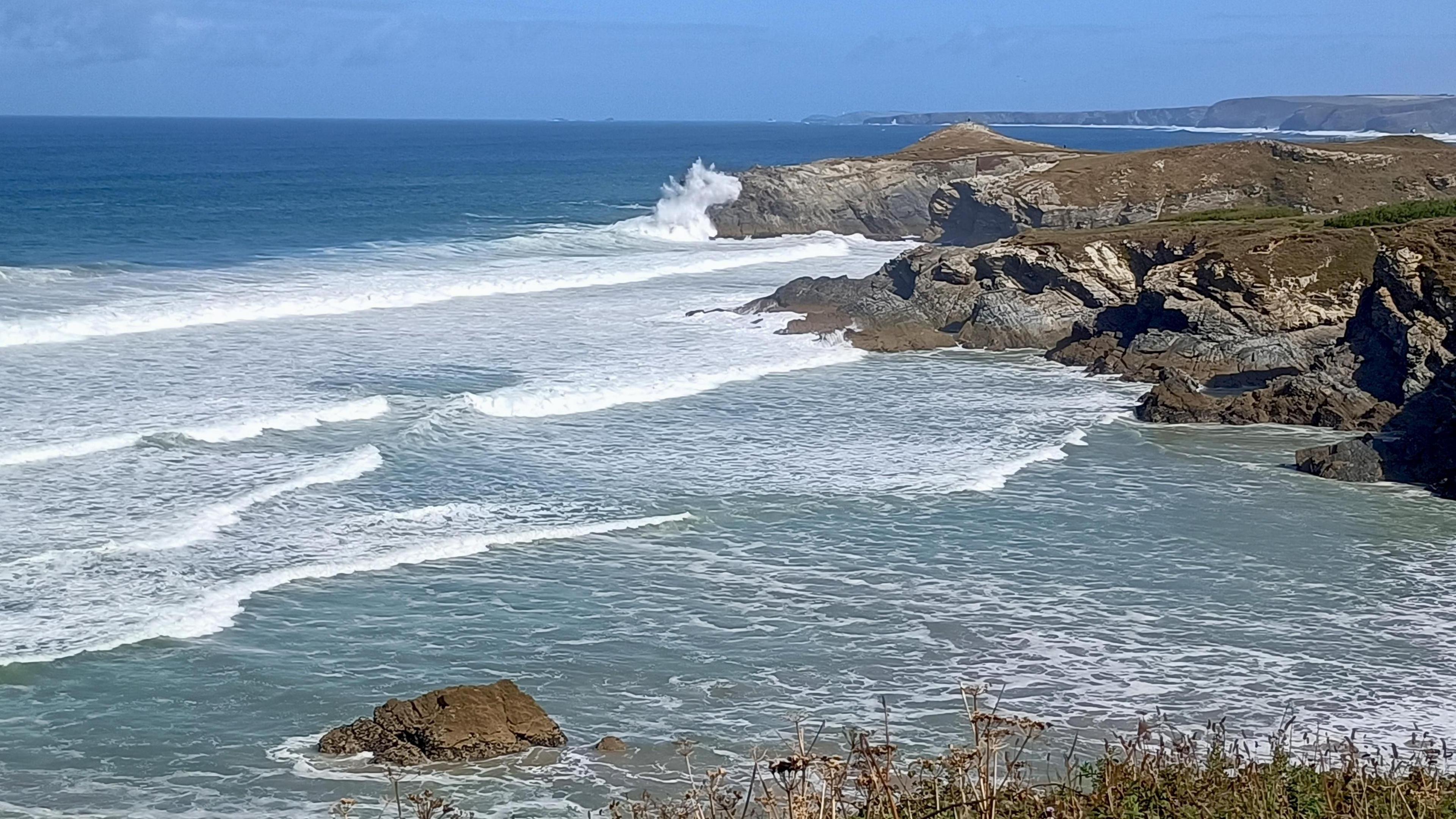 A small bay with a set of waves rolling in. They are white with wash and one is breaking on and splashing above the headland. Headlands can be seen further down the coast. It is a bright blue sky day.