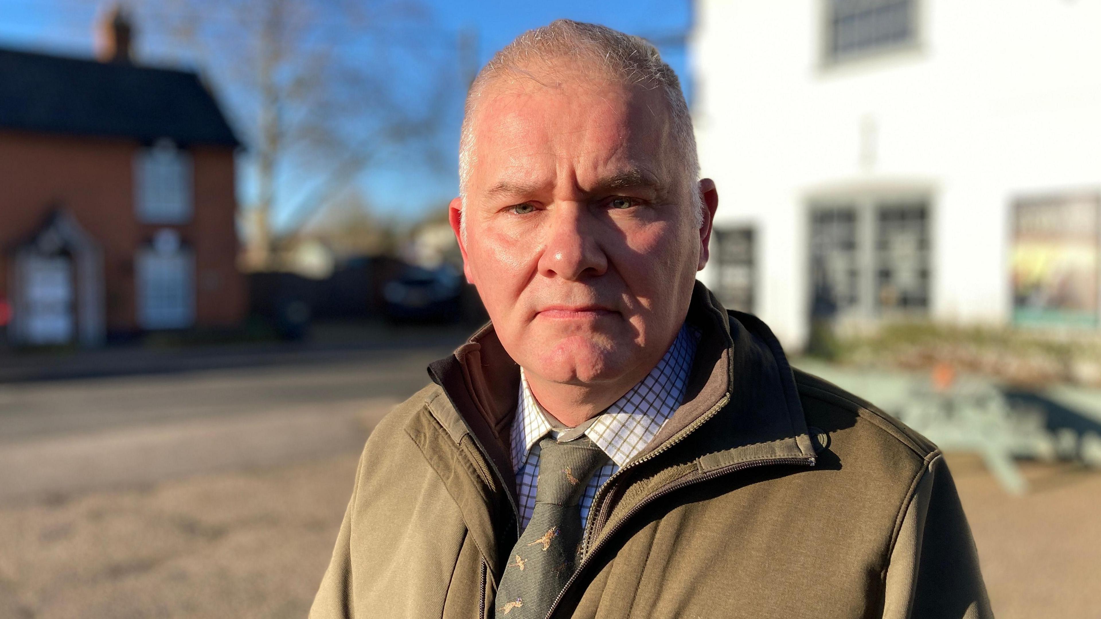 Independent councillor Clayton Hudson standing alongside a road. He has short grey hair, is cleanly shaven and is wearing a green coat over a shirt and tie.