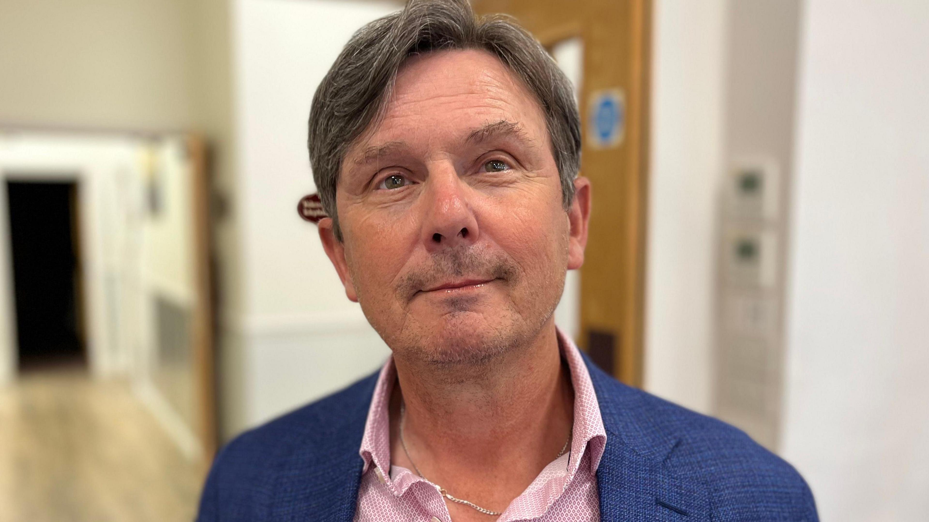 Deputy Andy Sloan - A man with black and grey hair. He is wearing a blue suit and a pink shirt with a silver chain underneath. 