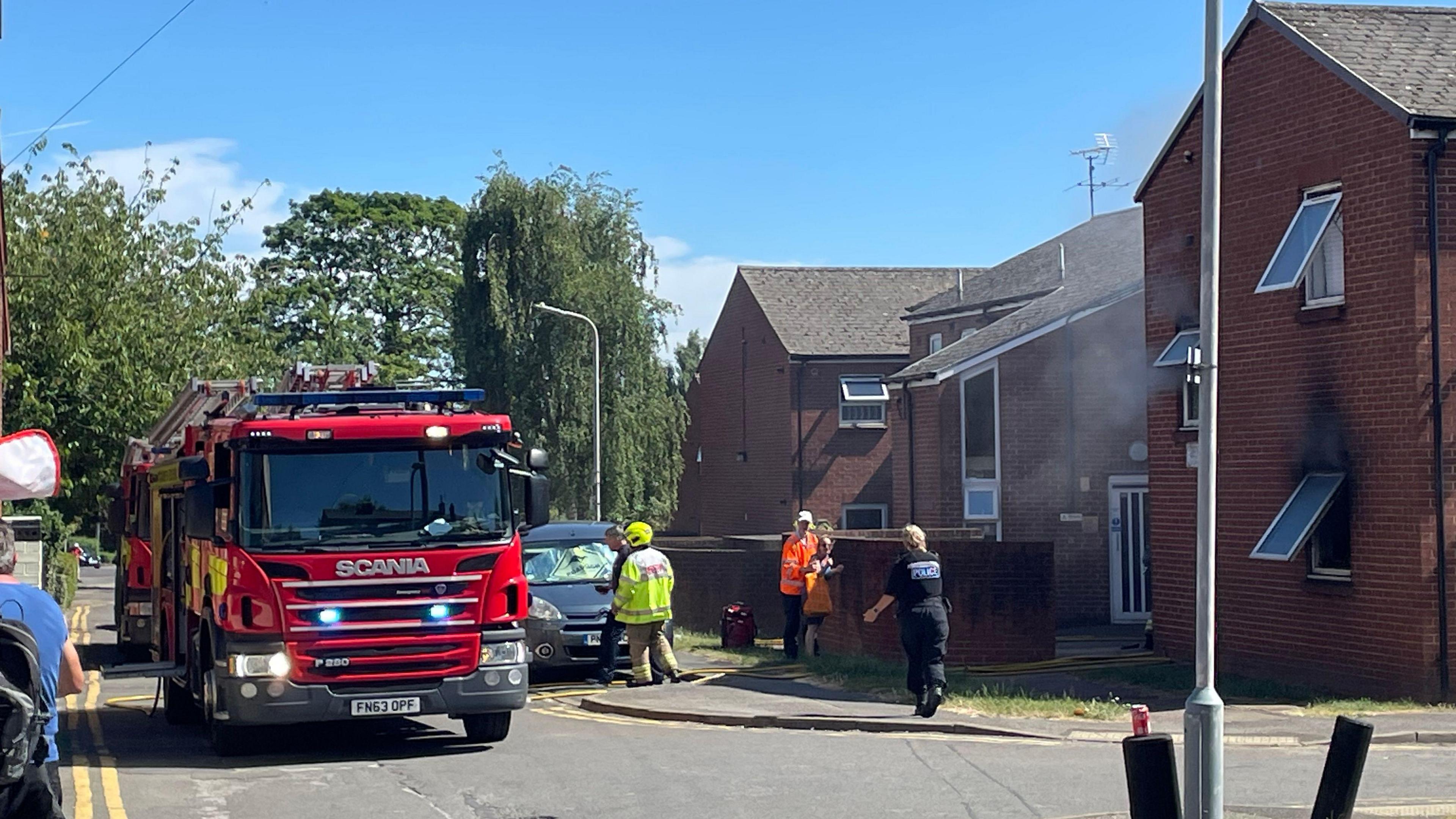 The scene of a flat fire, two fire engines are on the left of the image in the middle of the road, fire service staff and a police officer can be seen on the street. To the left of the image is a two-storey block of fats, some of the brickwork is burnt around a ground floor window