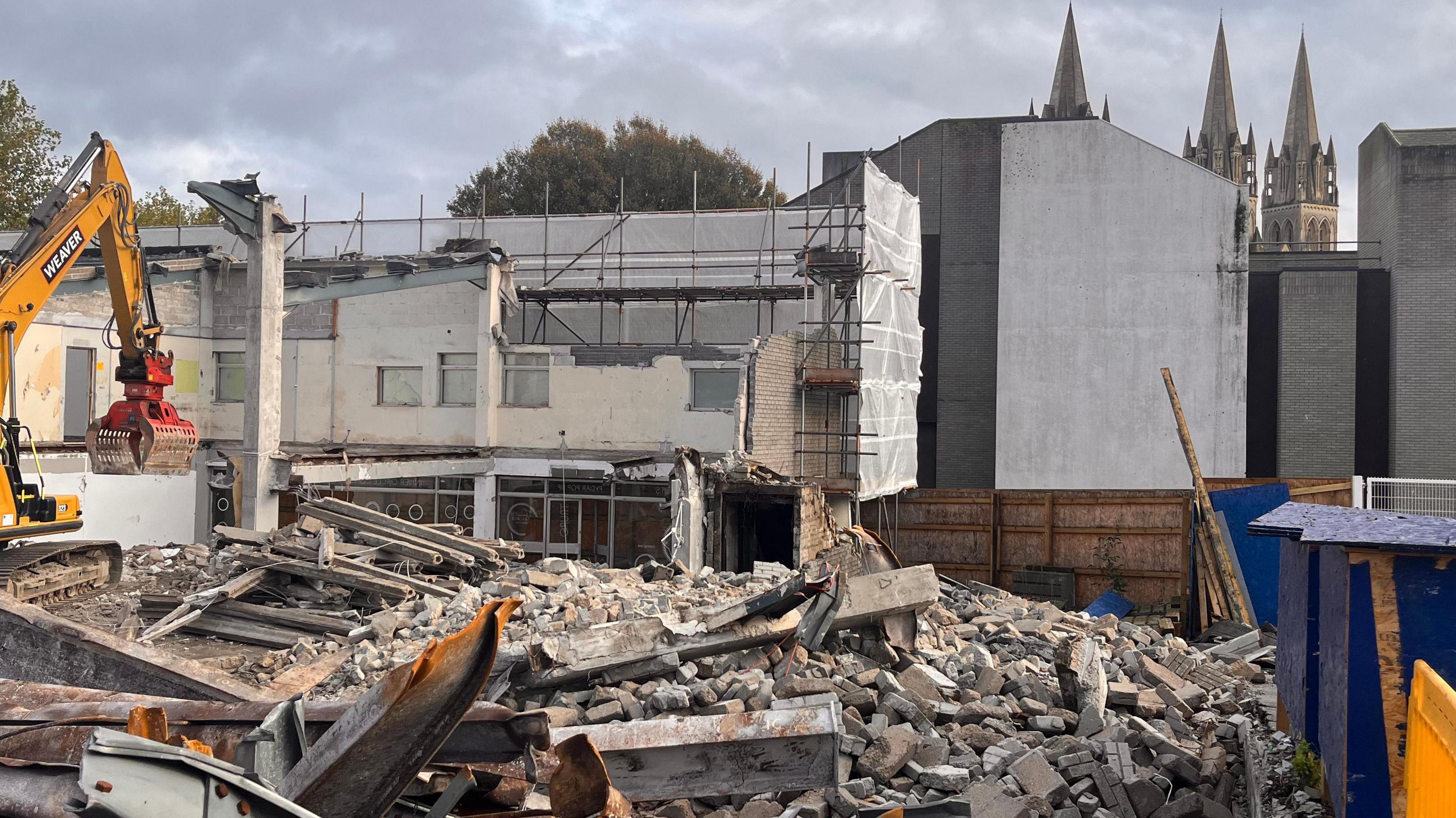 A Bulldozer demolishes old buildings in Truro , piles of rubble and are all around. The spires of Truro Cathedral can be seen in the background.