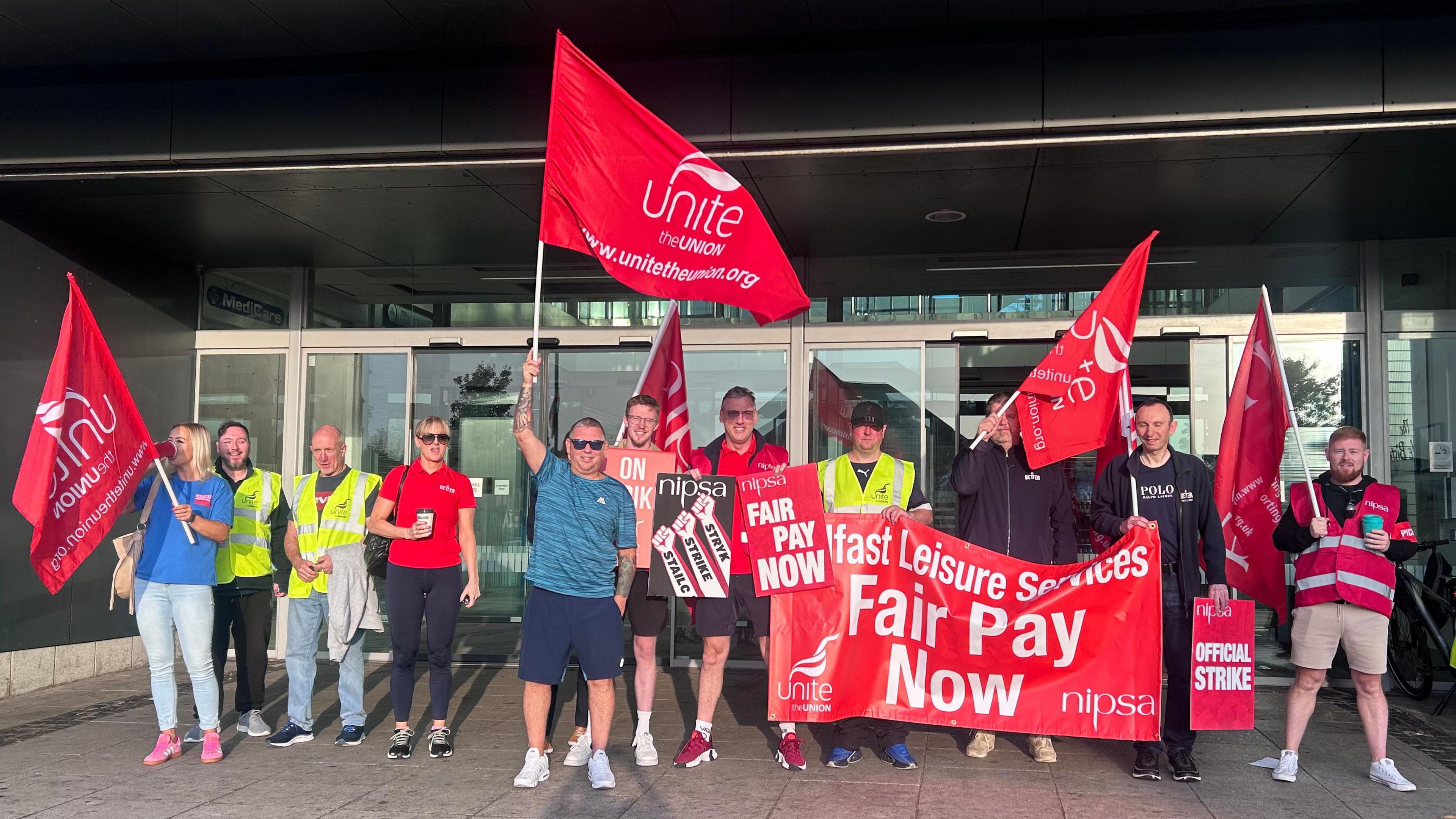 A group of people from Unite the Union on strike at the door of a leisure centre. They are wearing different coloured clothing and holding large red flags and a poster with the 'unite the union' and 'fair pay now' written on them. The doors behind them are glass and the ground is grey tiles.