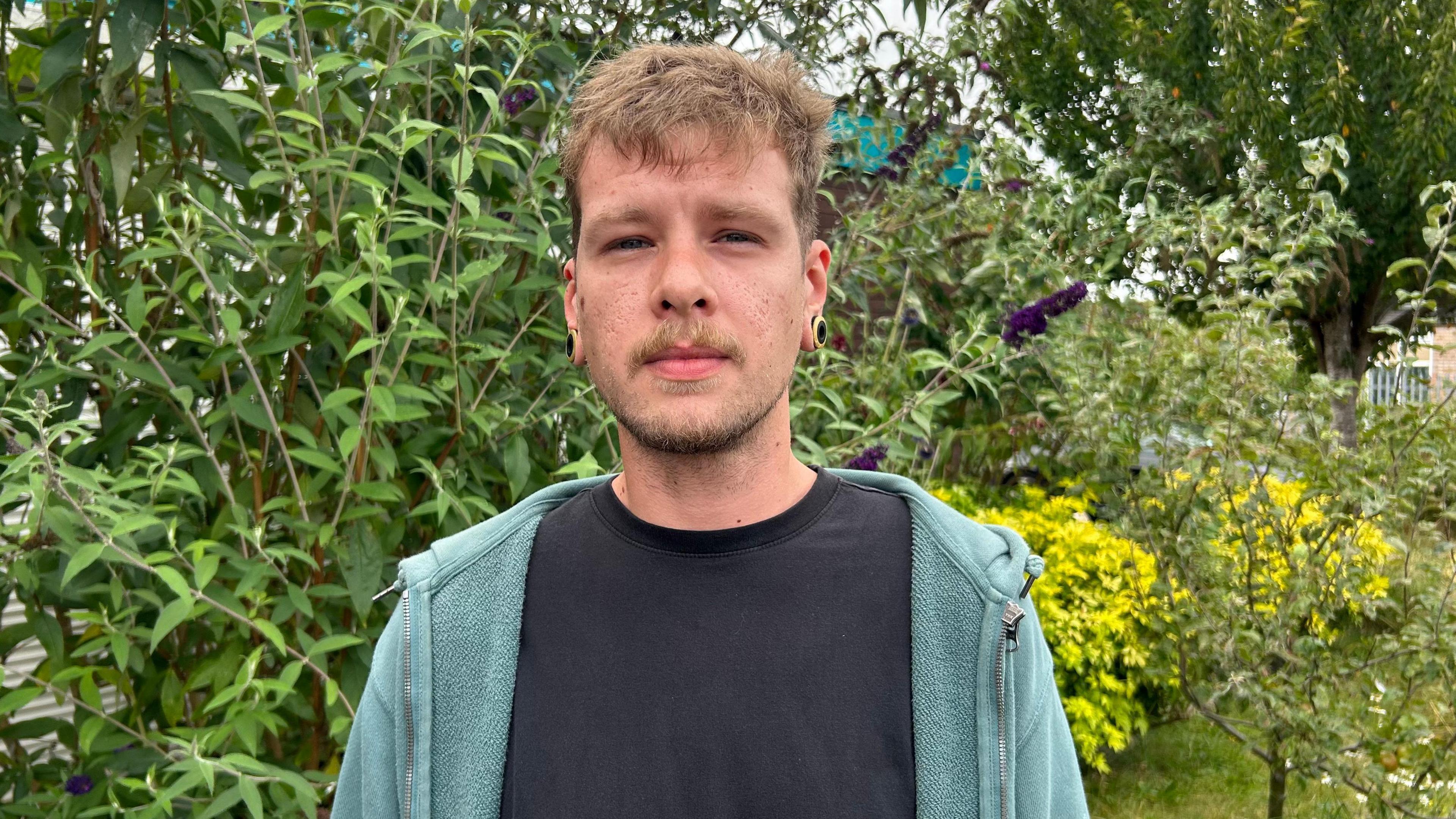 Connor Davey, a man in his 20s, wearing a black top and turquoise hoody, standing in front of a bush