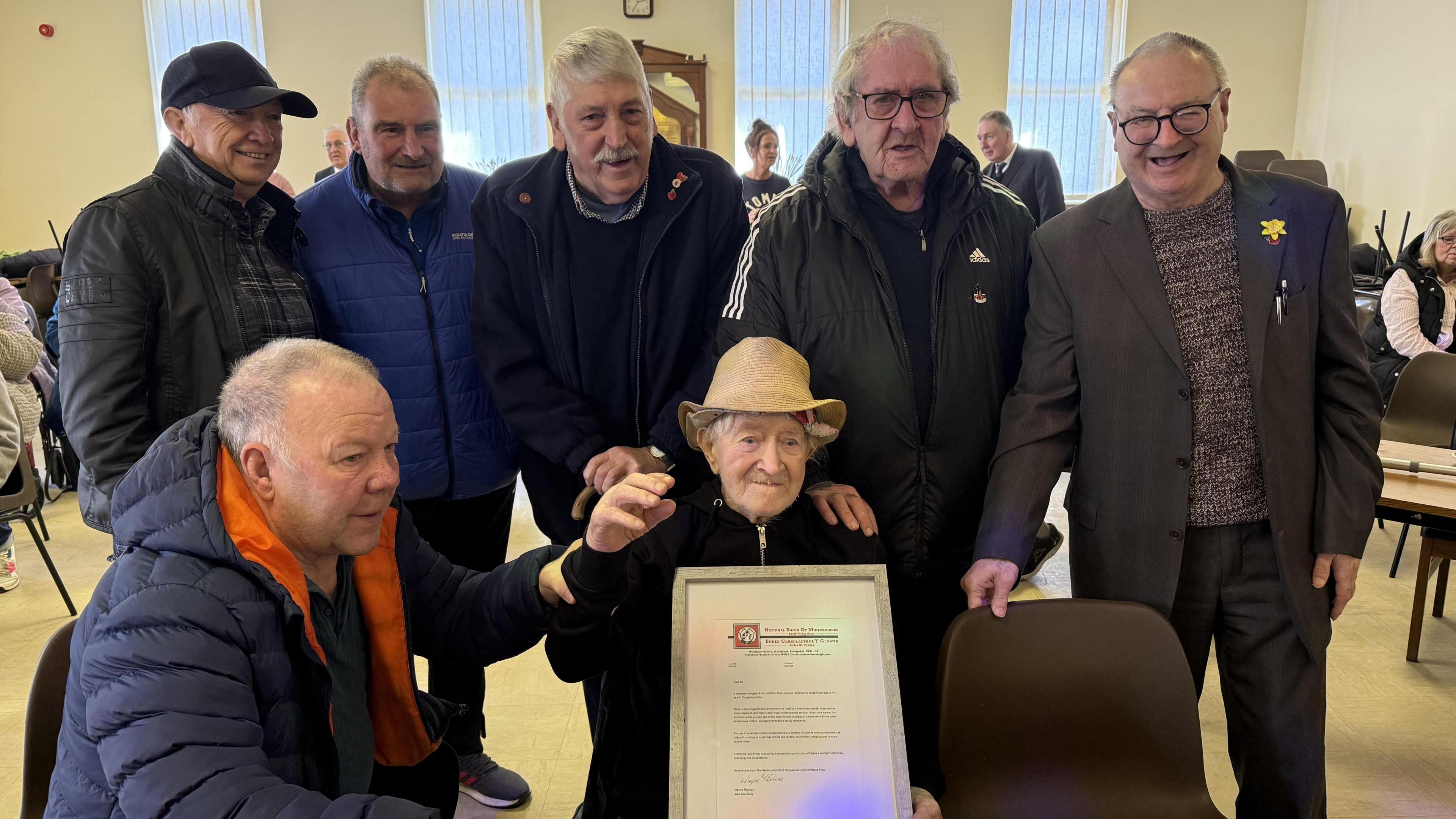 Ron "Ginger" Parsons sat in the middle of six men, five of whom are standing behind him. One man is sat next to Mr Parsons who is helping him hold up a certificate. Mr Parsons is wearing a straw hat and a black zip-up jumper. The other men are wearing black and navy coats. The group appear to be in a village hall.