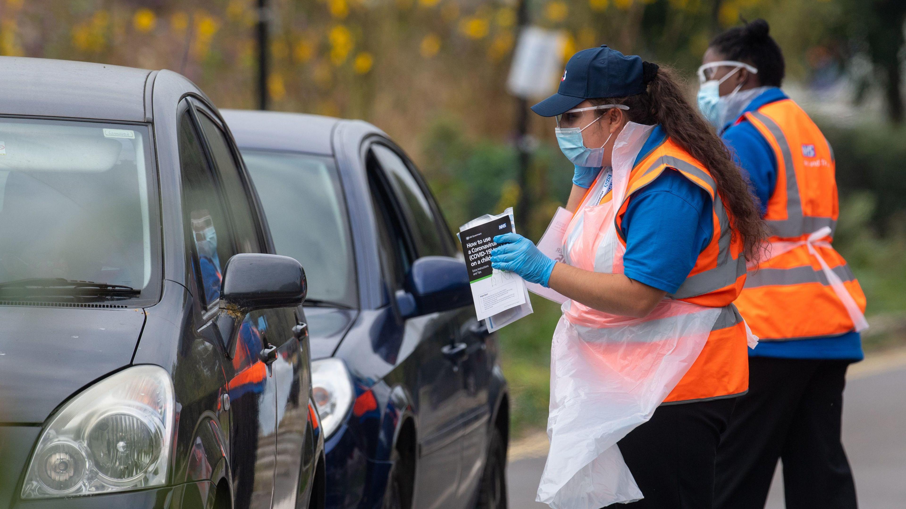 A drive-in testing site in Southwark, London on 16 September 2020. There are two dark cars queuing to pick up testing kits from staff wearing masks, aprons and gloves.