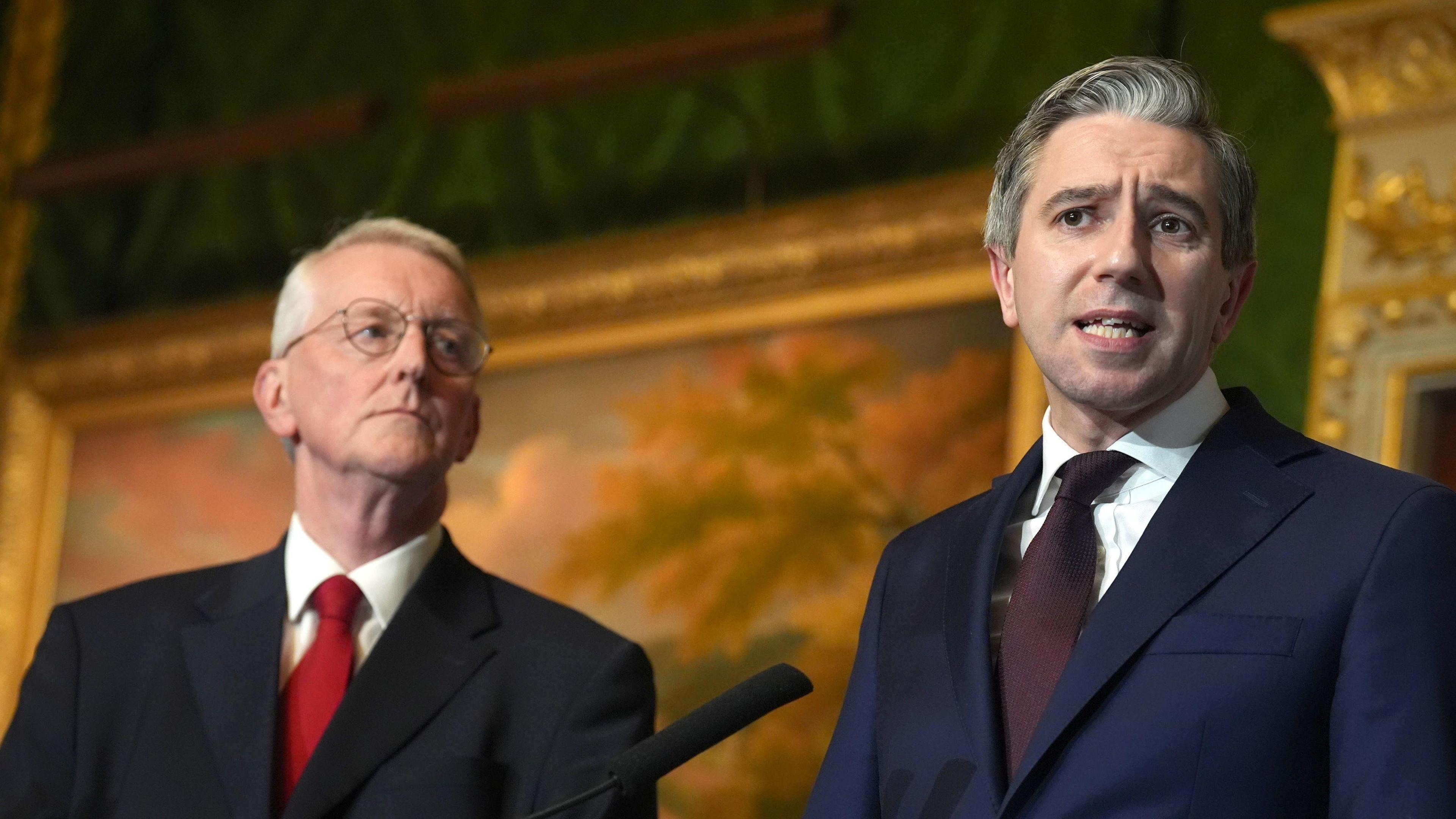 Hilary Benn and Simon Harris standing beside each other. Benn is wearing glasses, a black suit, white shirt and red tie. He is looking at Harris, who is wearing a navy suit, burgundy tie and white shirt.