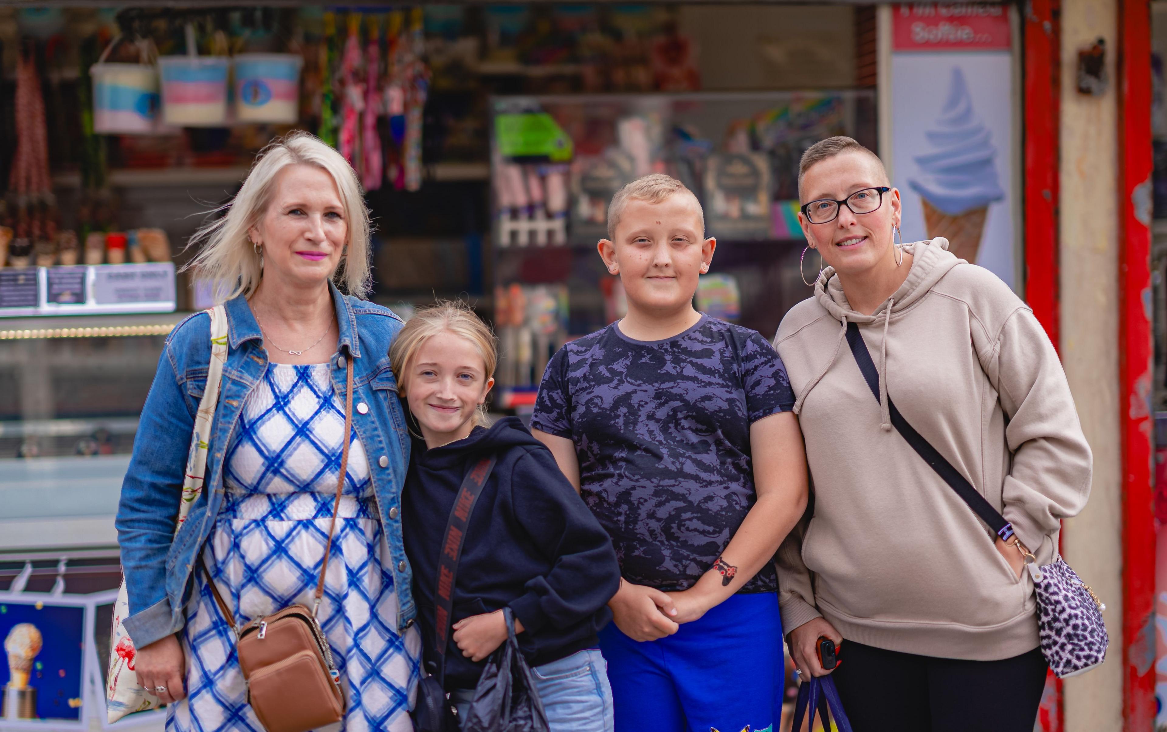 A family posing for a photograph in front of an ice cream stall. A woman in her 50s with blond hair and a blue dress with her arm around a young girl in a dark hoody. A boy in blue t-shirt and shorts and a woman with short hair and a brown hood stand beside them.