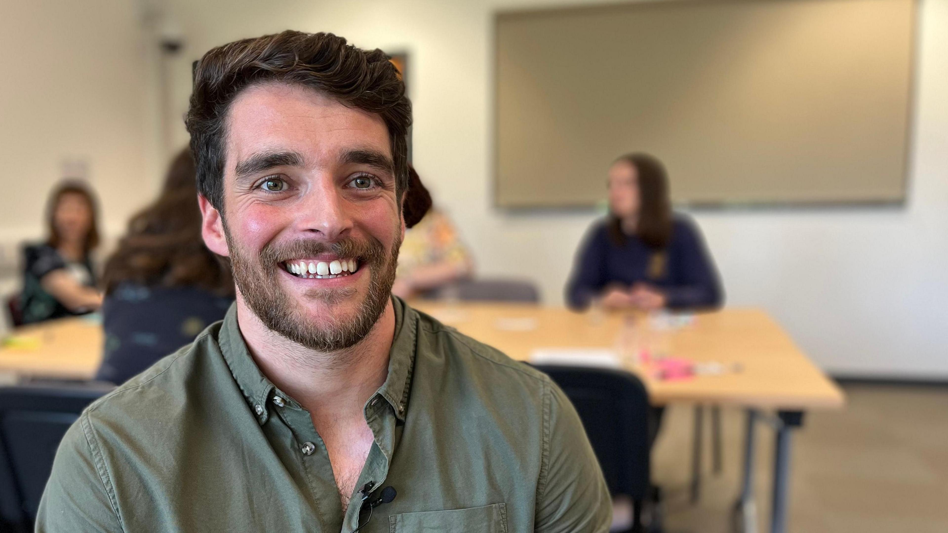 A man with dark hair is wearing a khaki green shirt and smiling at the camera. Behind him is a large desk with a number of women sat around it talking. The man has a beard and dark eyes.