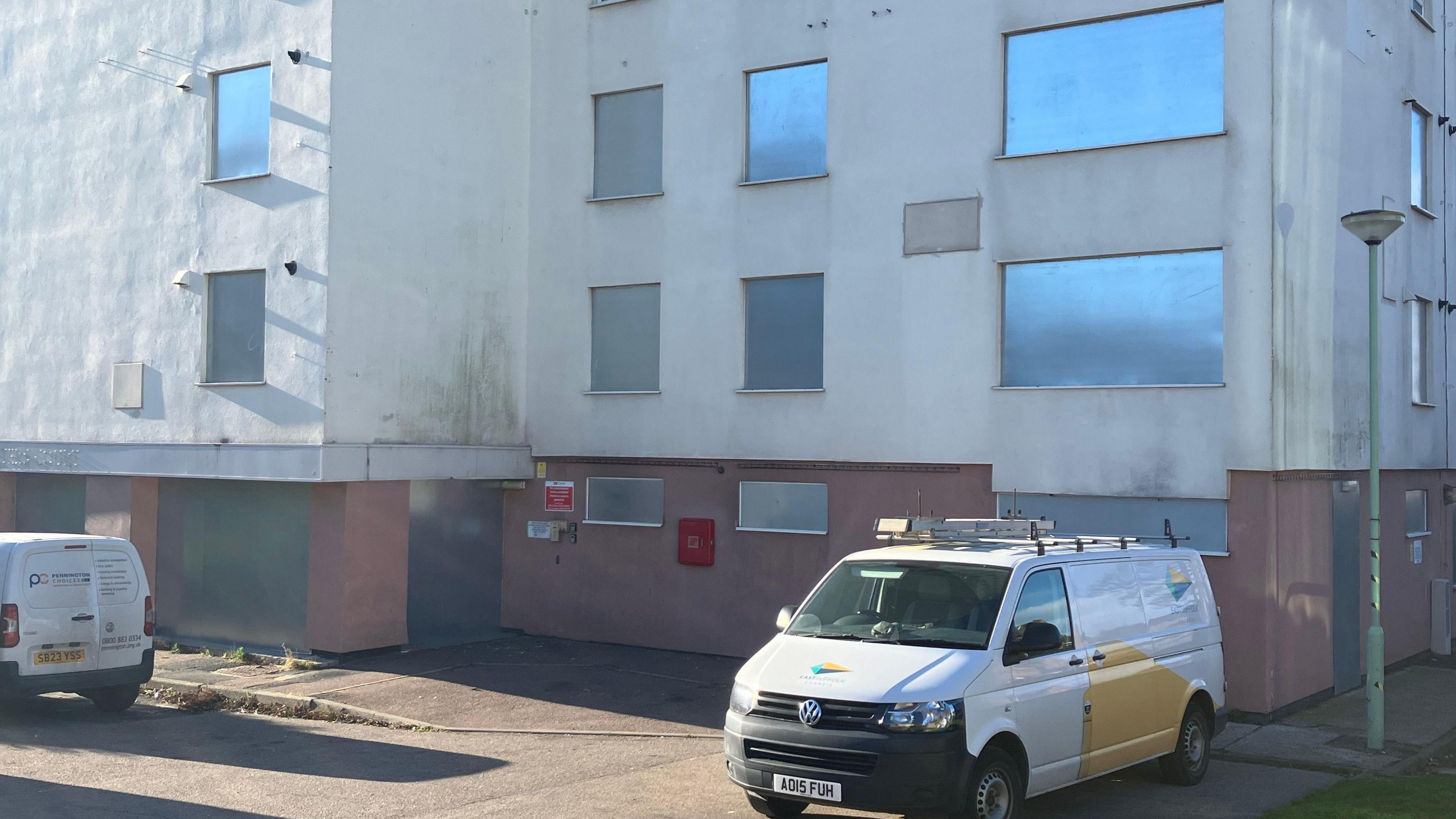 Silver metal covers all the windows in the first two storeys of the white tower block and two council vans can be seen parked in the driveway