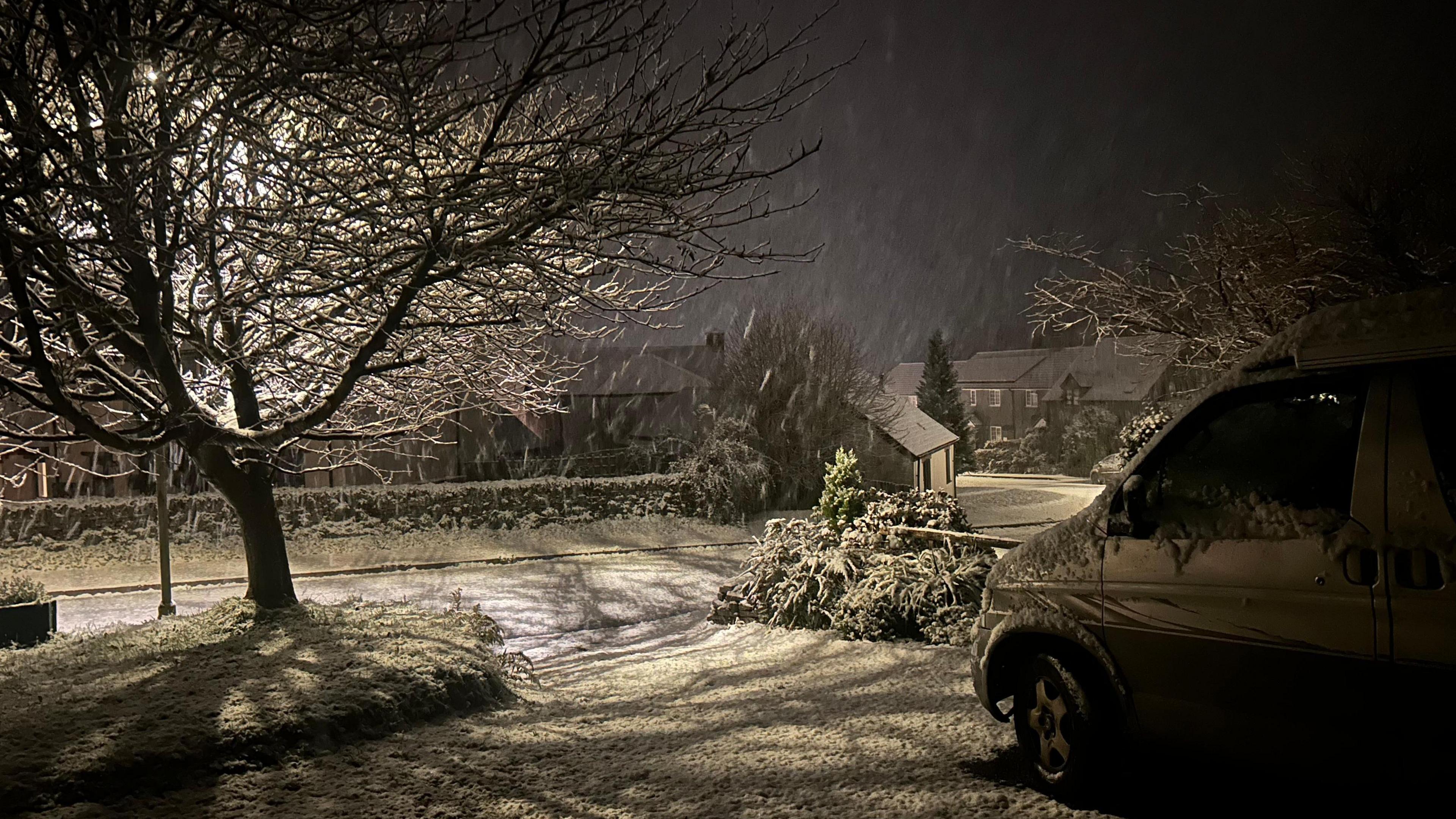 Heavy snow is falling down in an area of Cornwall. A van is parked in a driveway. It is dark. A tree is being lit up by a lampost.