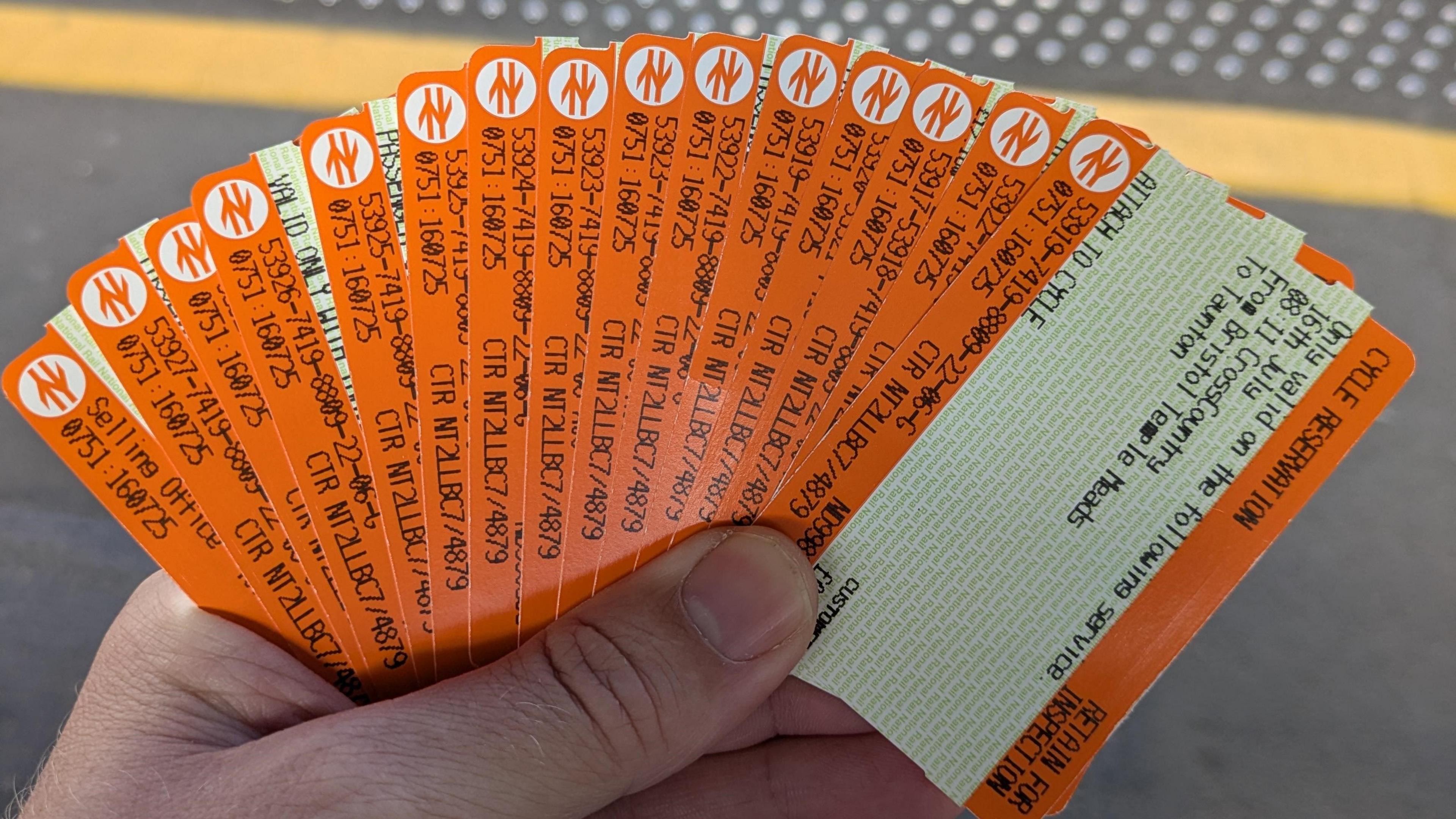 A man standing on a train platform holding a large fan of 15 orange train tickets in his left hand.
