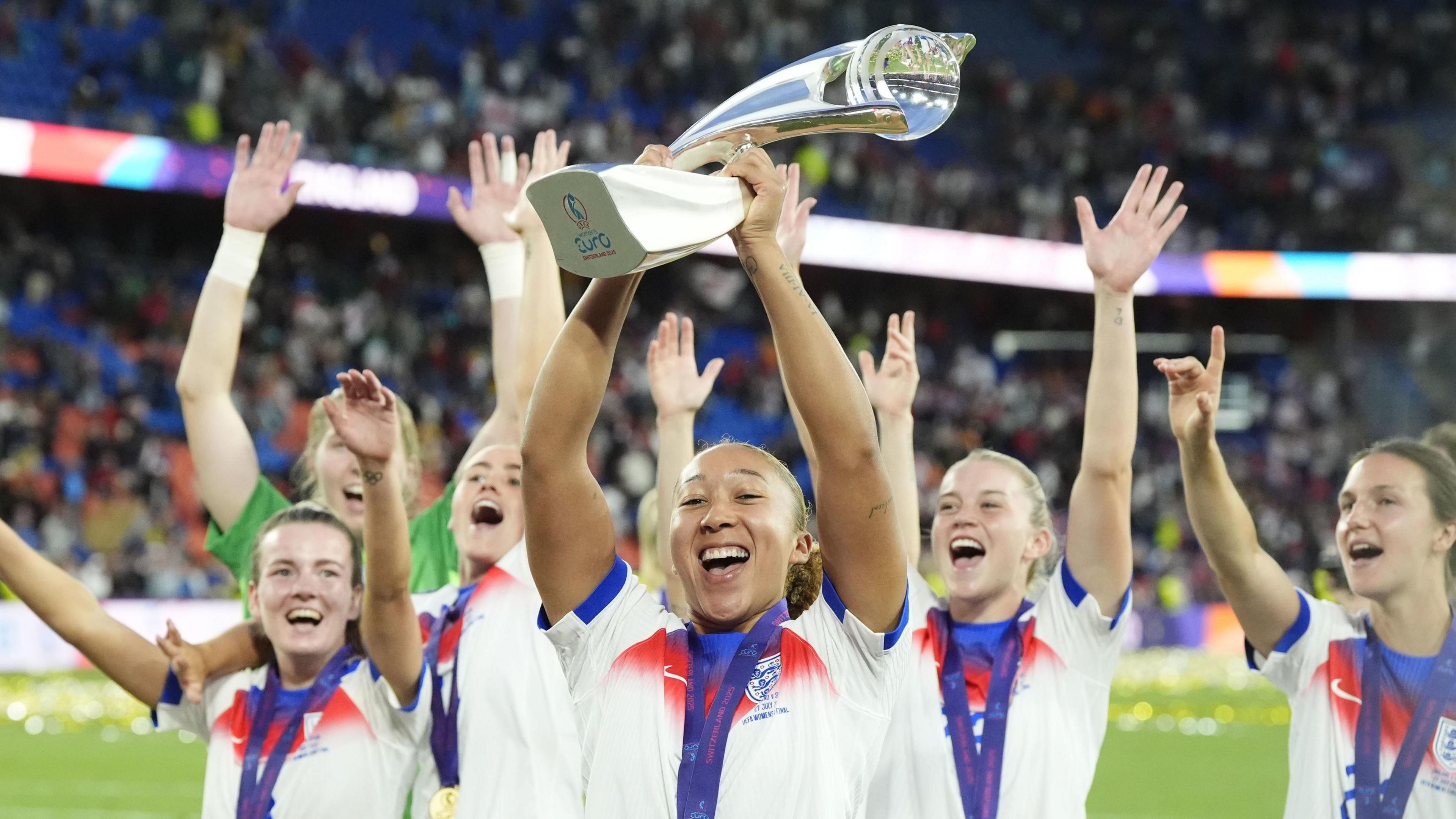 England's Lauren James celebrates with the trophy after winning the Euro 2025 final. She is joined on the pitch by five other players with their arms in the air.