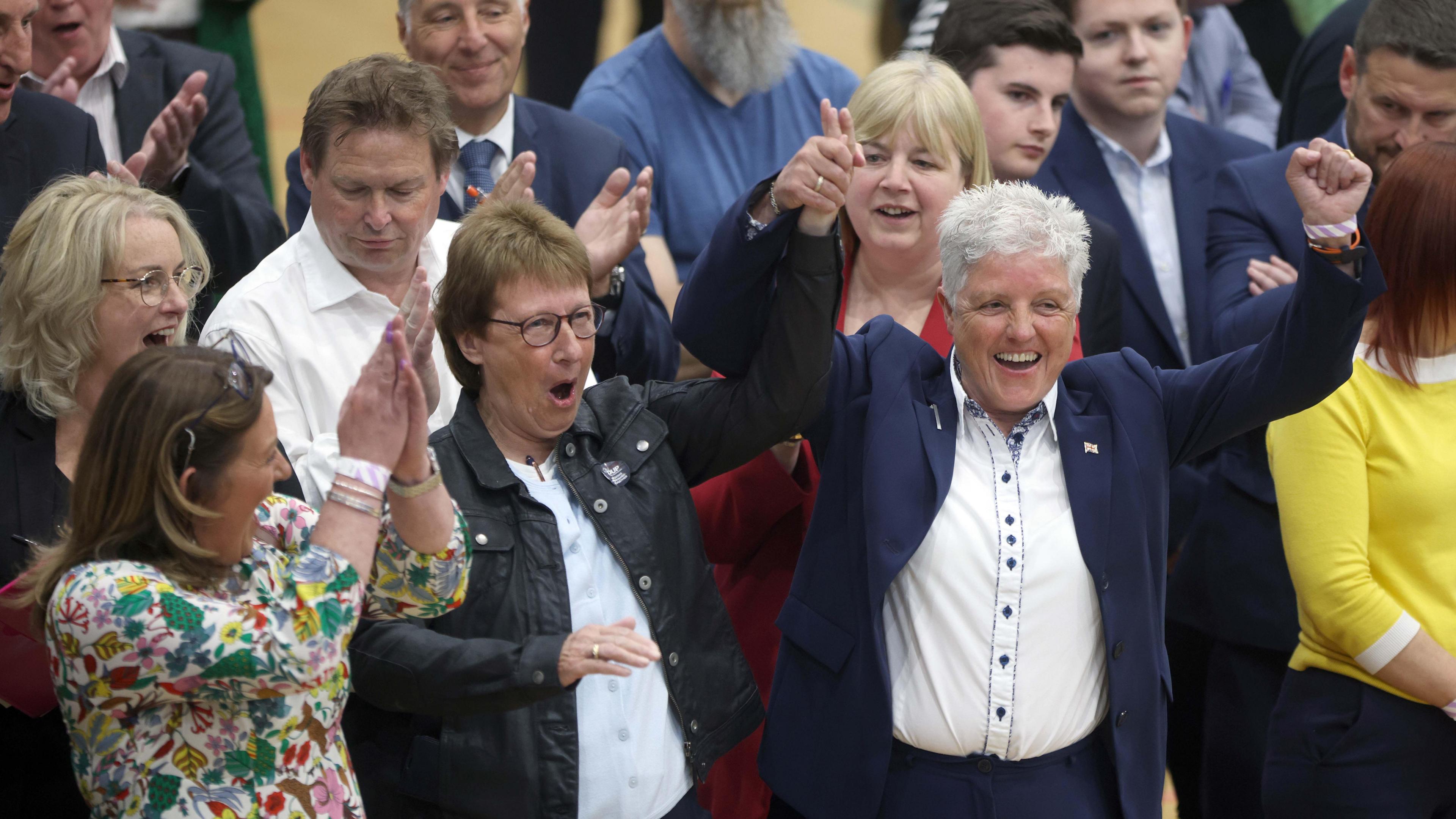 Alison Bennington at the 2023 NI local elections count being congratulated by cheering supporters. She has her arms aloft and several women and men beside her are smiling and applauding.