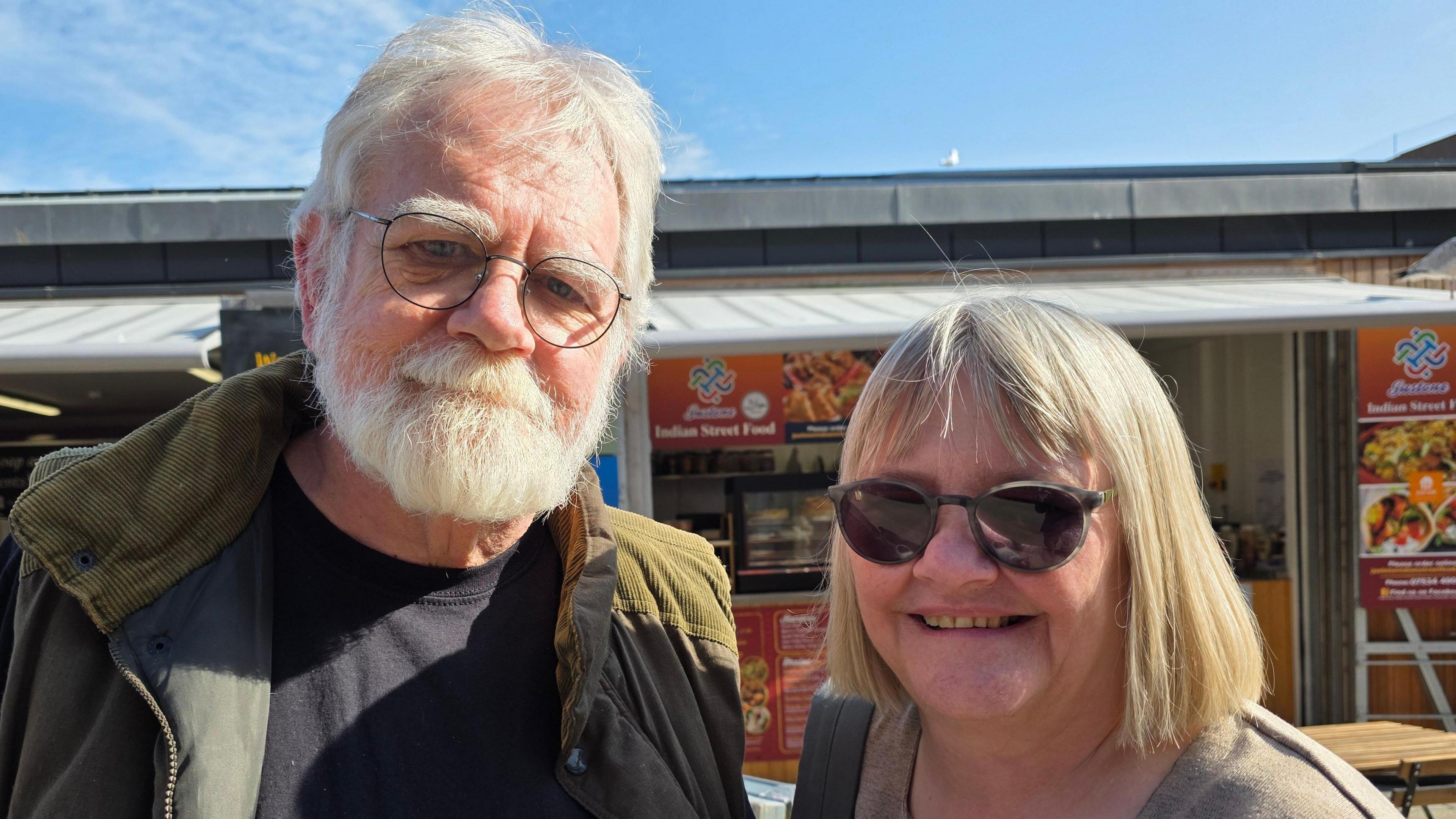 David and Ruth Woollard-Kingston are standing in front of a market stall. He is wearing glasses and a green coat over a black t-shirt. She is wearing shades.