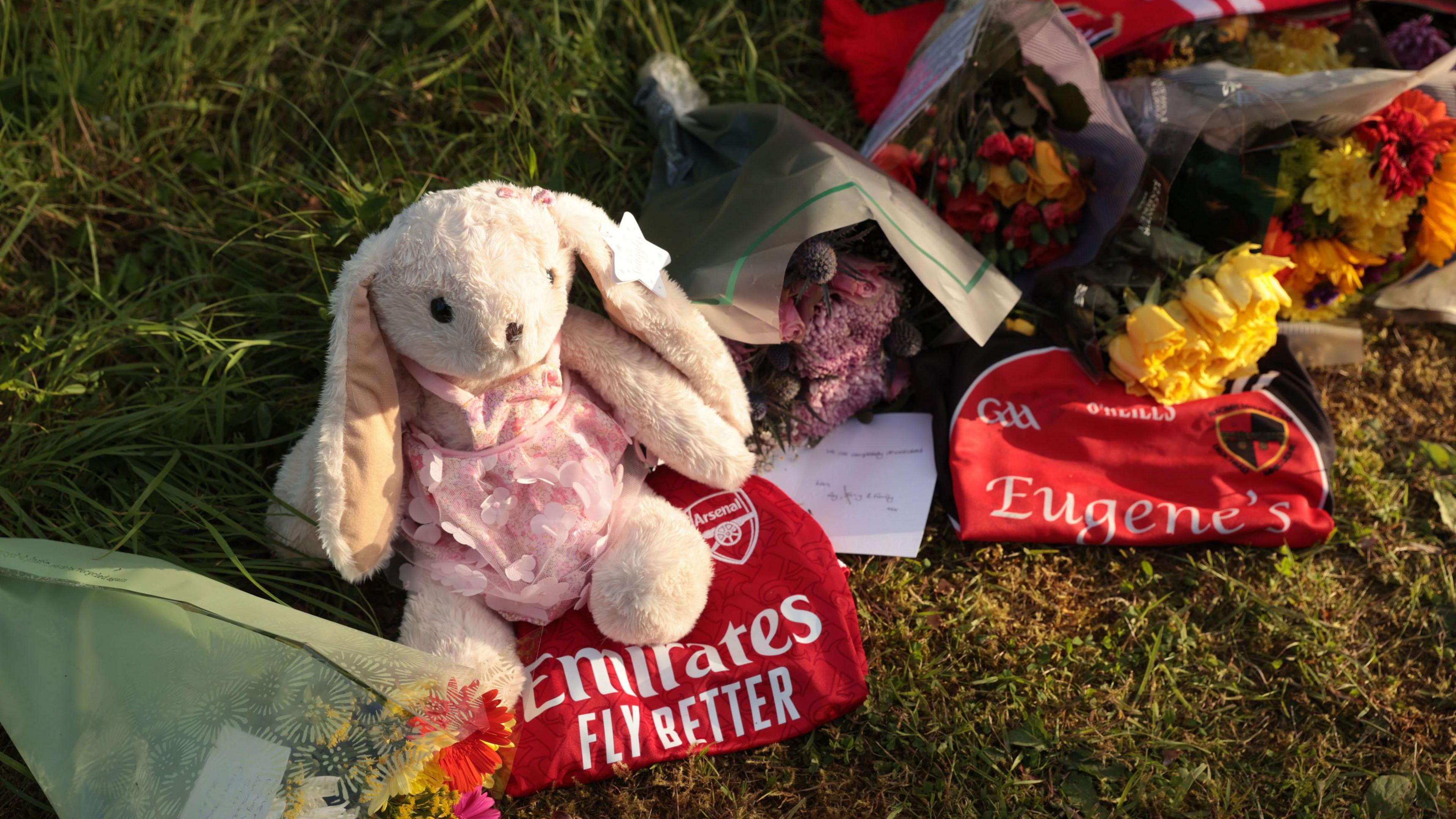 A rabbit teddy, flowers, an arsenal scarf and jersey and a GAA jersey are lying in grass