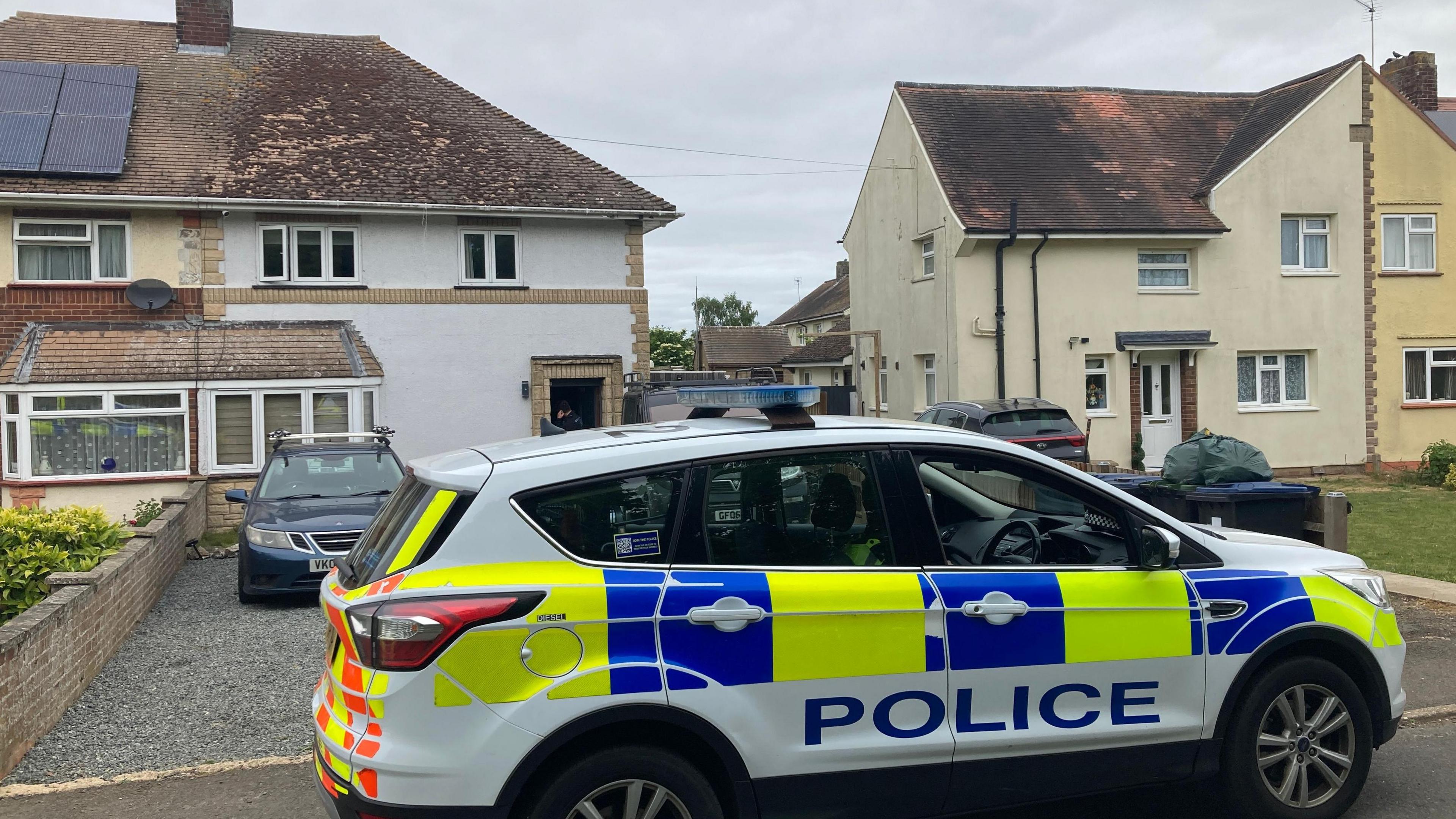 A police car outside a semi-detached white house. It is blocking in the paved over garden of the house.