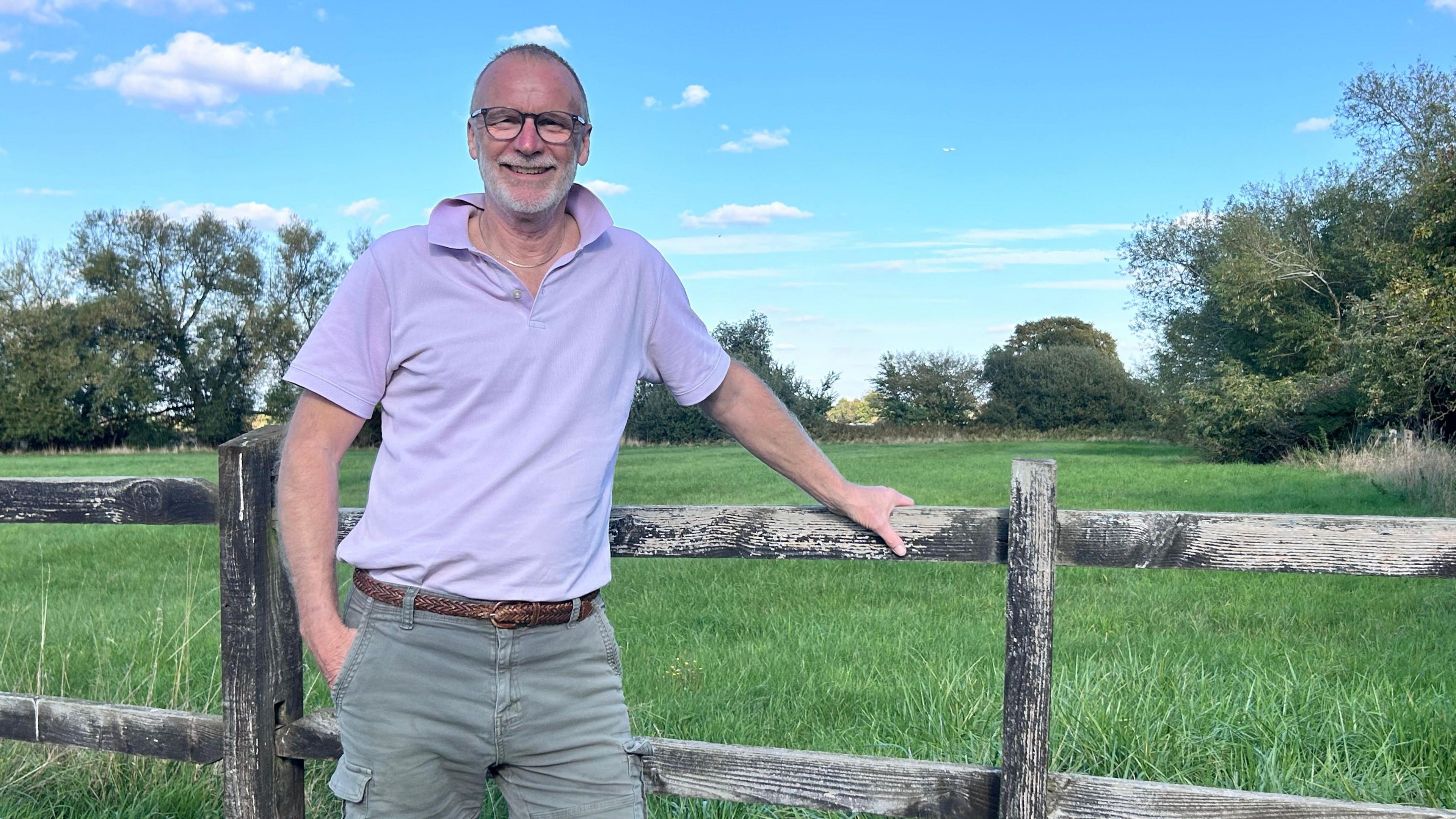 A man, who is wearing glasses, is wearing a light purple polo shirt and khaki shorts. He is standing next to a wooden fence and in front of a field. 