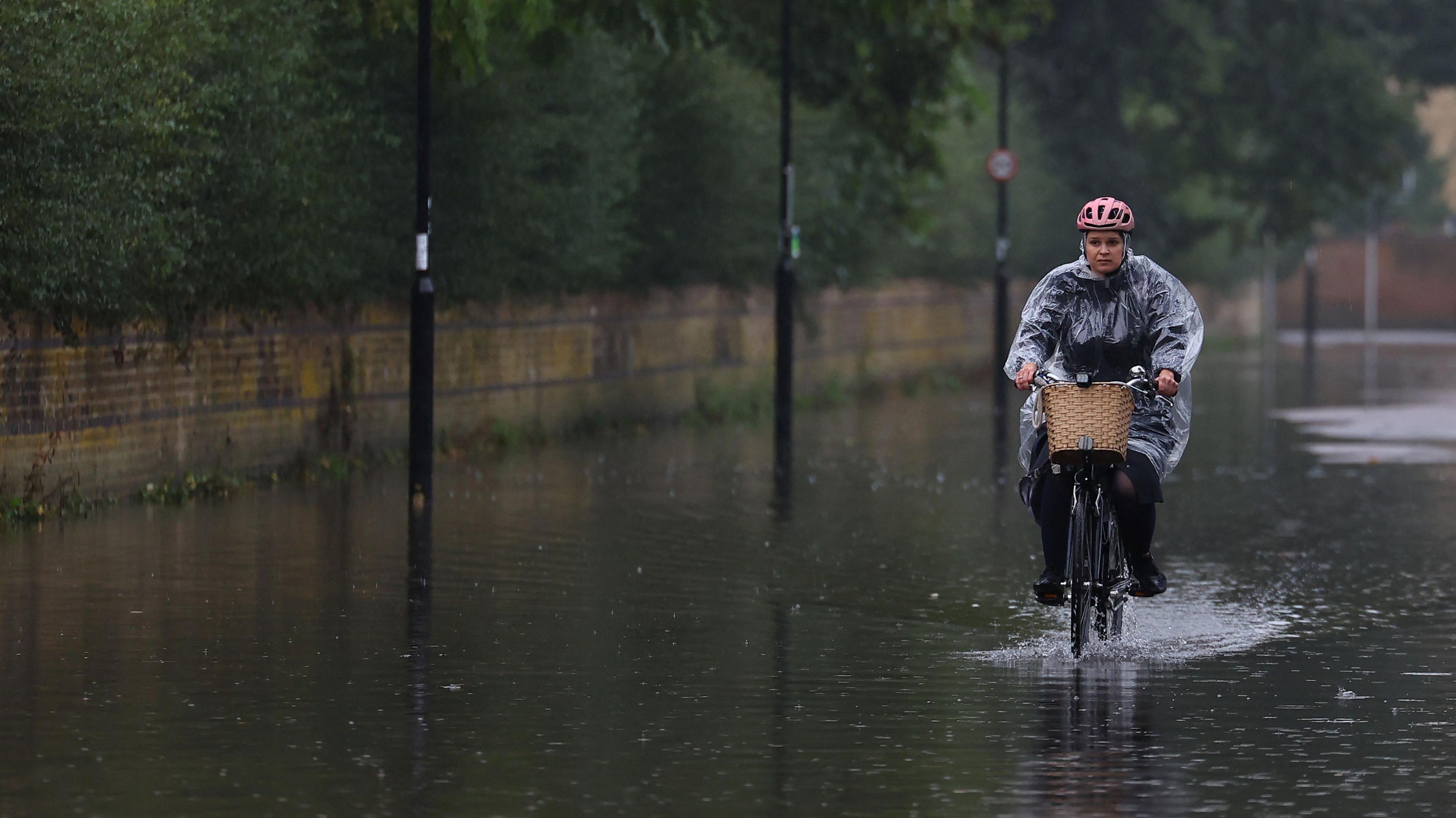 A female cyclist pedals though flood water on a road. The woman wears a clear plastic rain poncho, black clothes and a pink helmet. Her bicycle has a wicker basket. Water has filled the road and surrounded lampposts. 