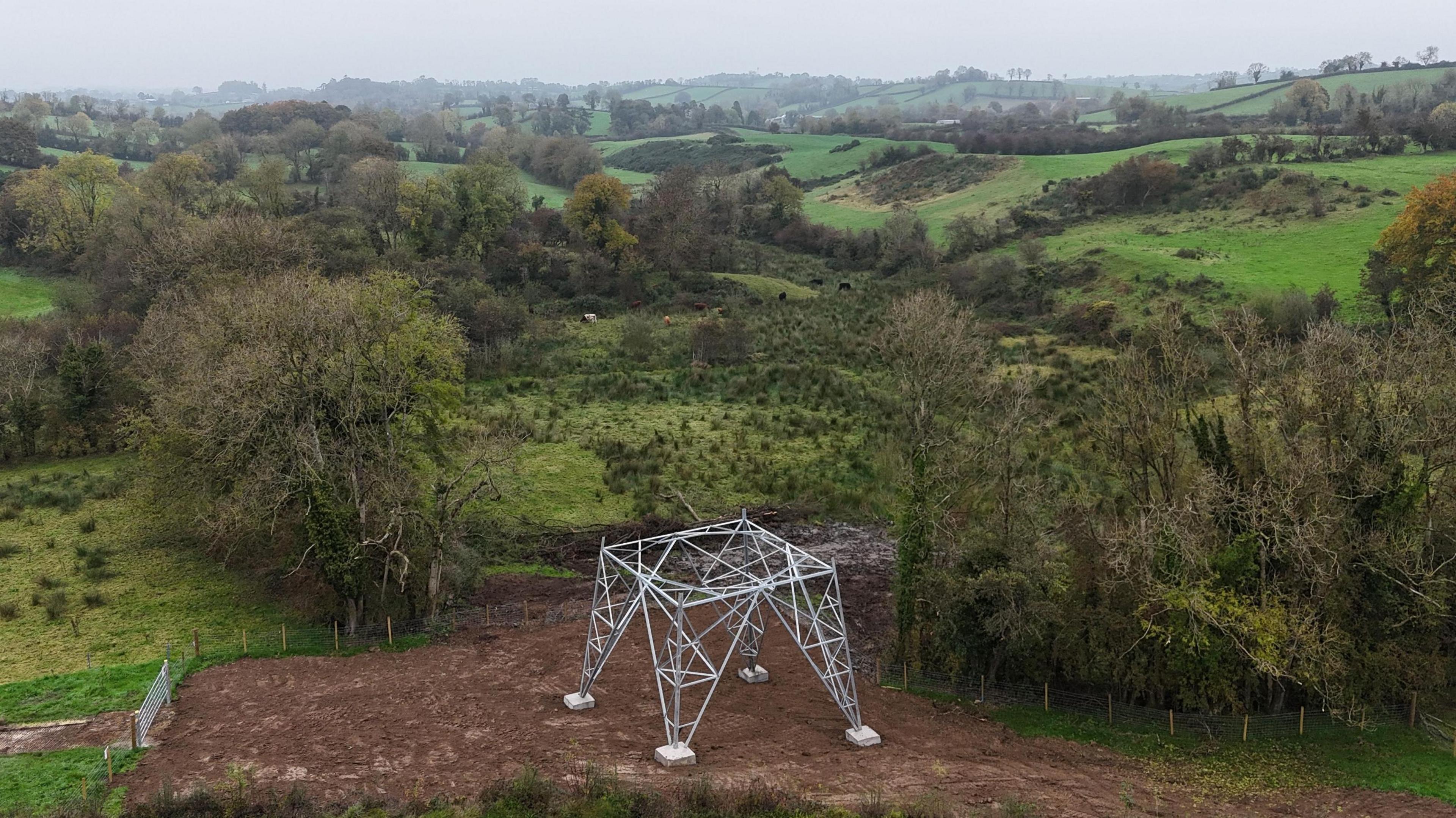A partially built steel pylon standing in a rural landscape surrounded by uncultivated dark green fields, trees and low hills rolling into the horizon.