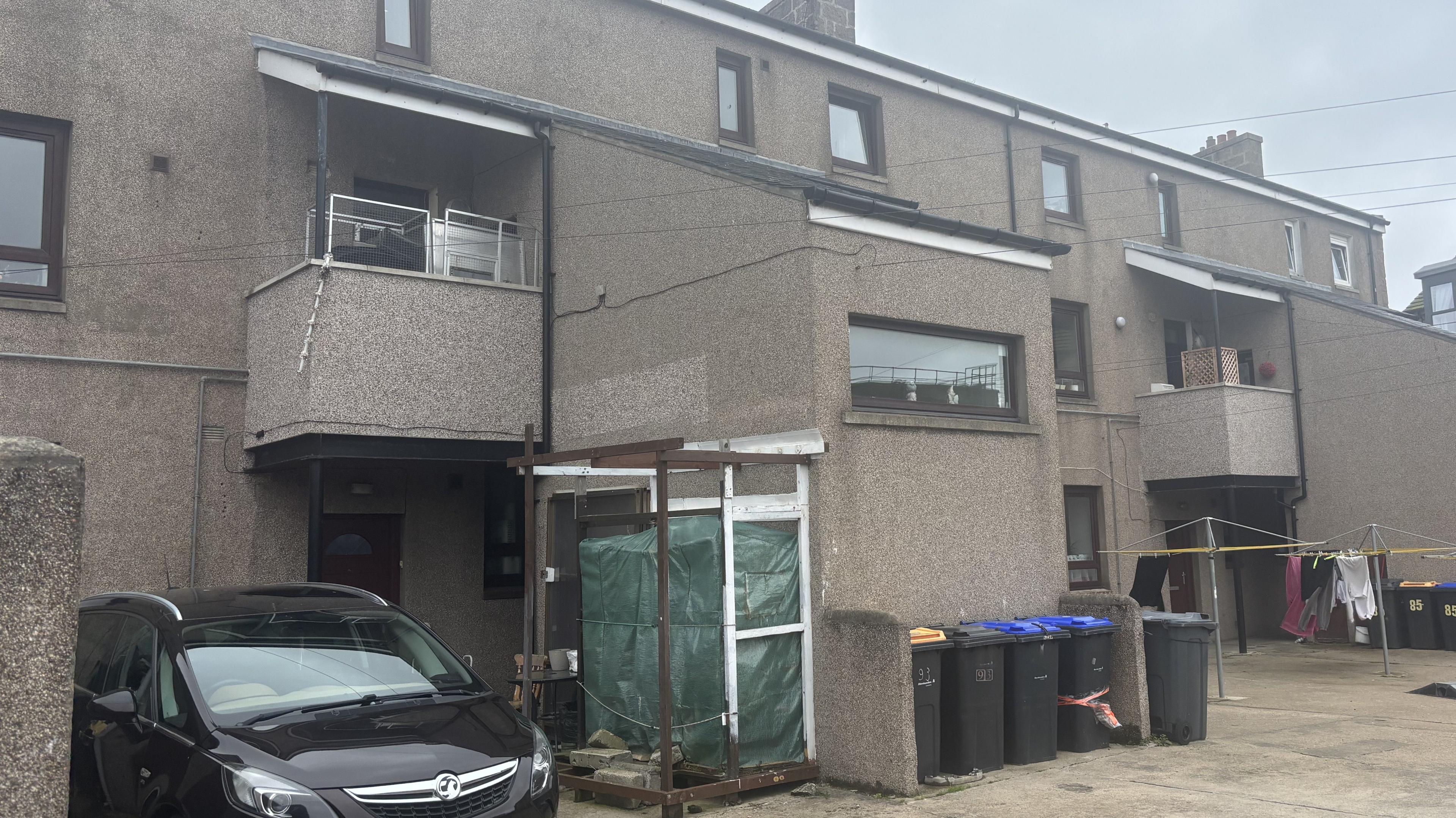 Rear view of a grey block of flats. A black Vauxhall car is parked in the yard. Five grey wheelie bins are also visible as well as a washing line.