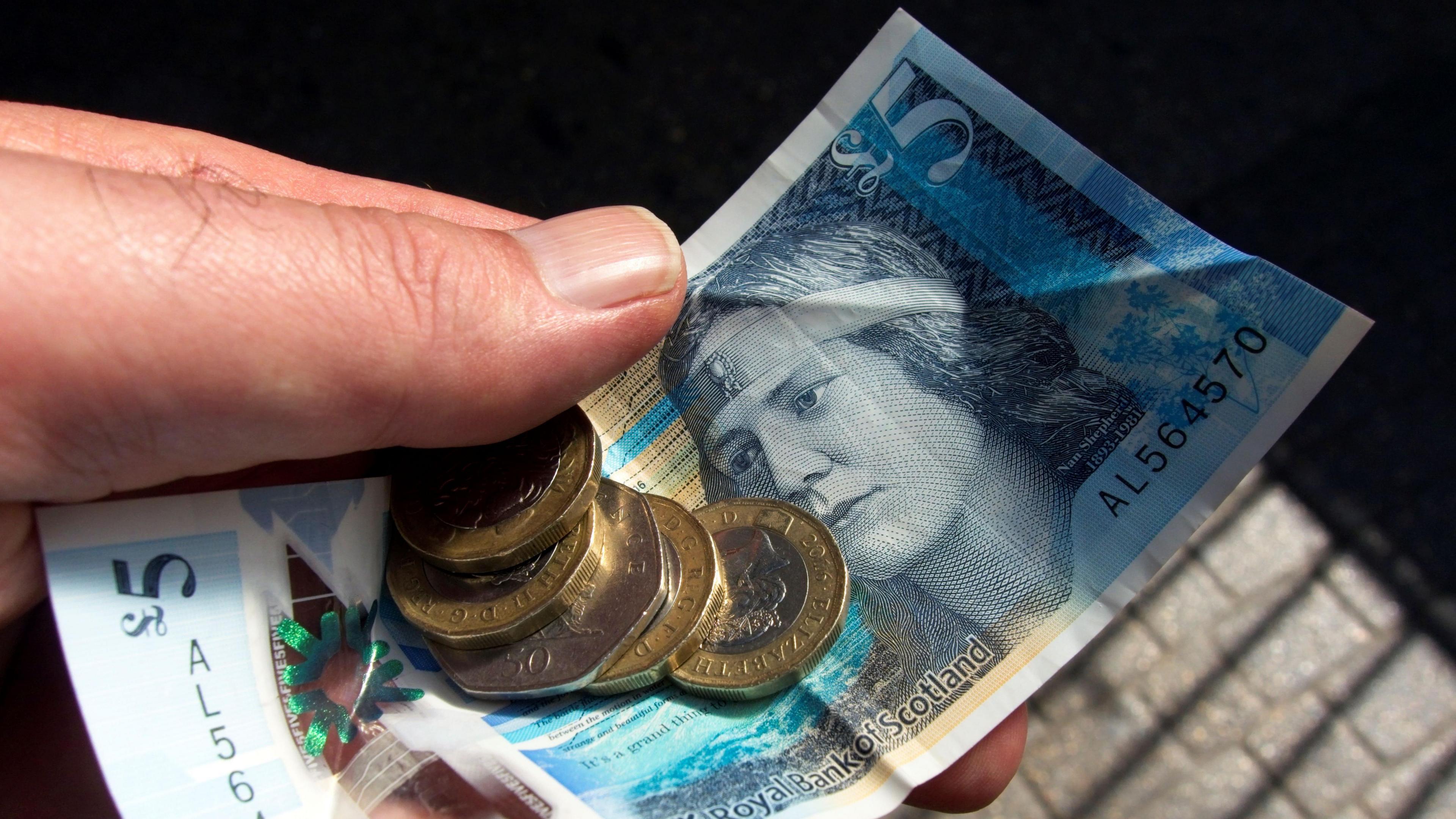 A hand holding a five pound note and several coins