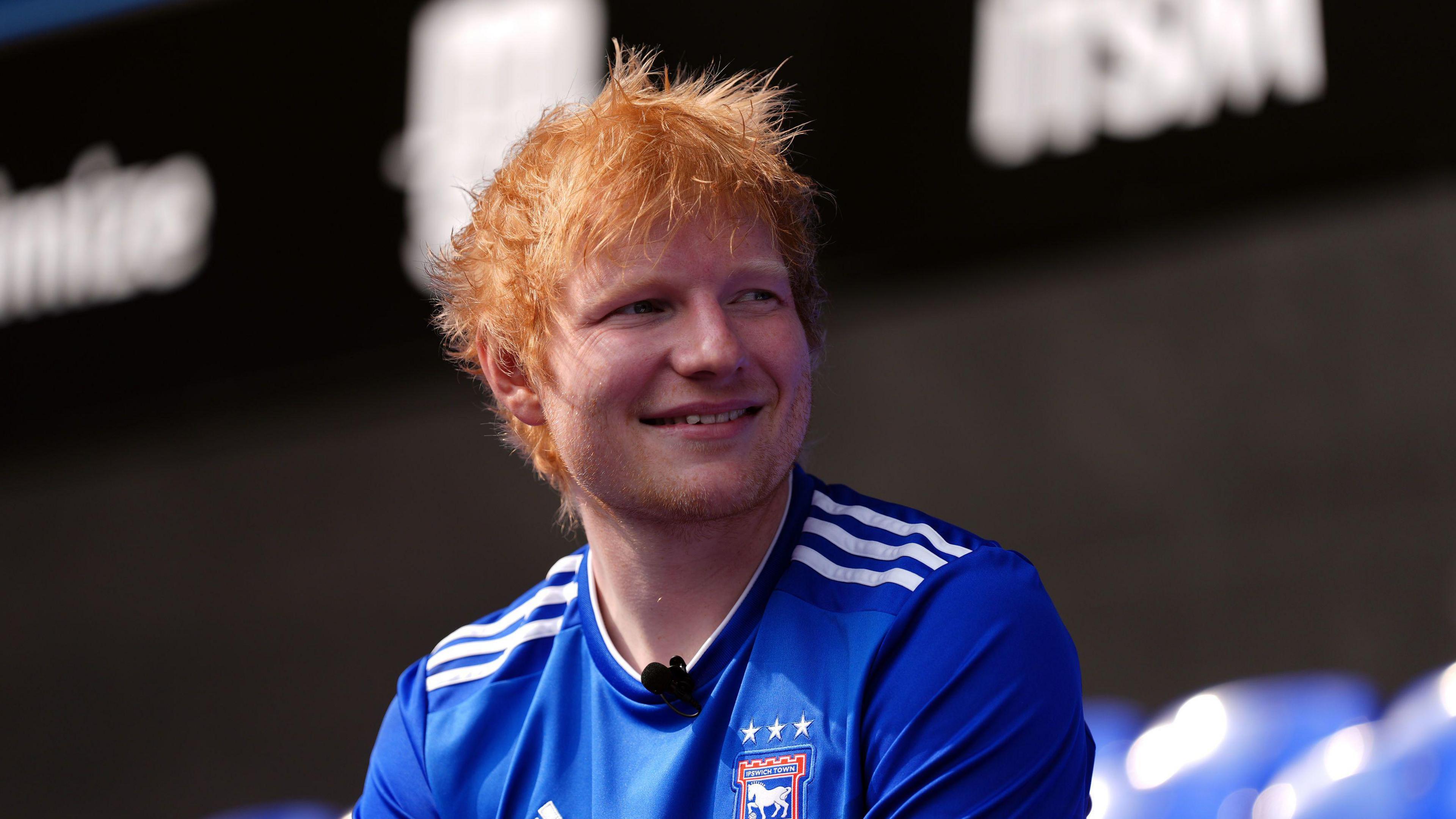 Ed Sheeran is pictured inside Portman Road stadium in Ipswich. He is looking away from the camera and smiling. He is wearing the blue, home Ipswich Town football shirt.