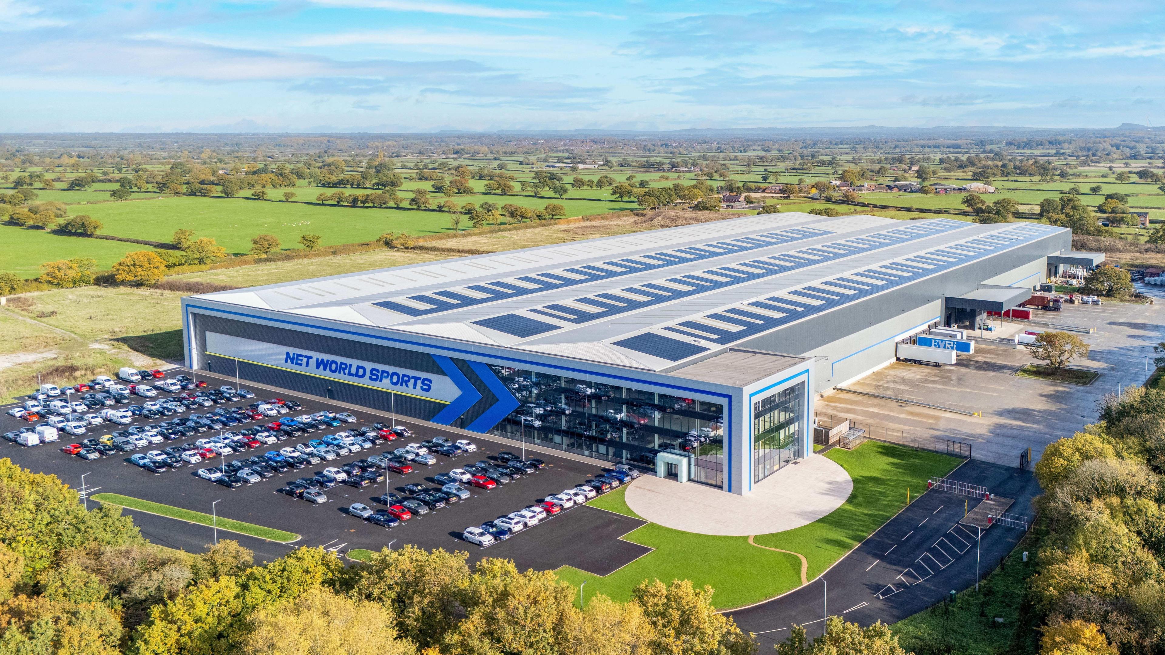 A large modern industrial building with “NET WORLD SPORTS” displayed prominently on the front in bold blue lettering. The structure has a glass-fronted office section on one side and an extensive warehouse area behind. Several rows of parked cars fill the adjacent car park, and a few lorries are positioned near loading bays. The site is surrounded by greenery, with open fields stretching into the distance under a partly cloudy sky.