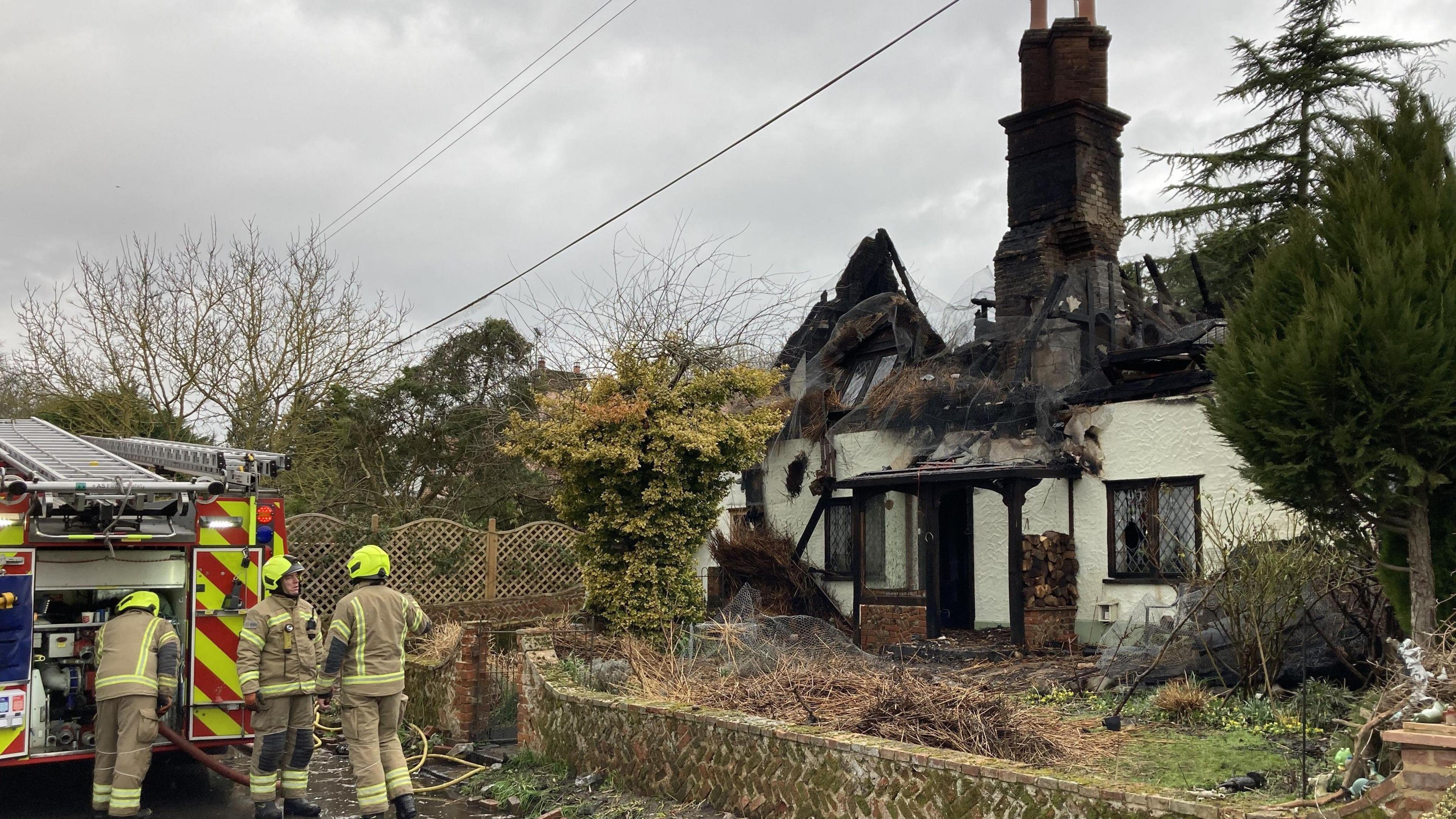 Firefighters stand near their fire engine outside the cottage. They wear firefighting uniforms and safety hats.