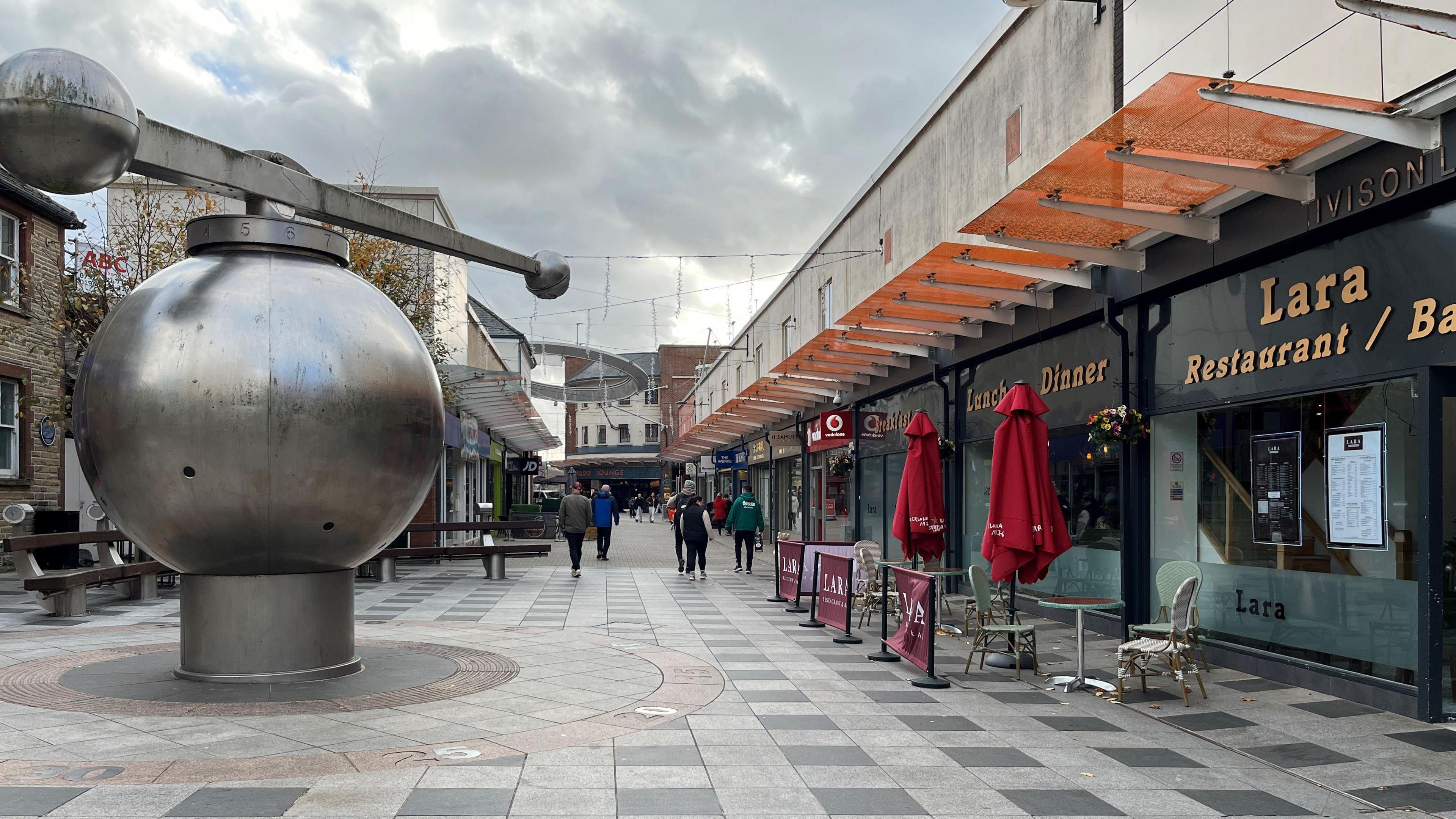 A general view of the shopping area in Workington town centre. The pedestrian area has a large metal sculpture in the middle, with shops either side and people walking down the street.