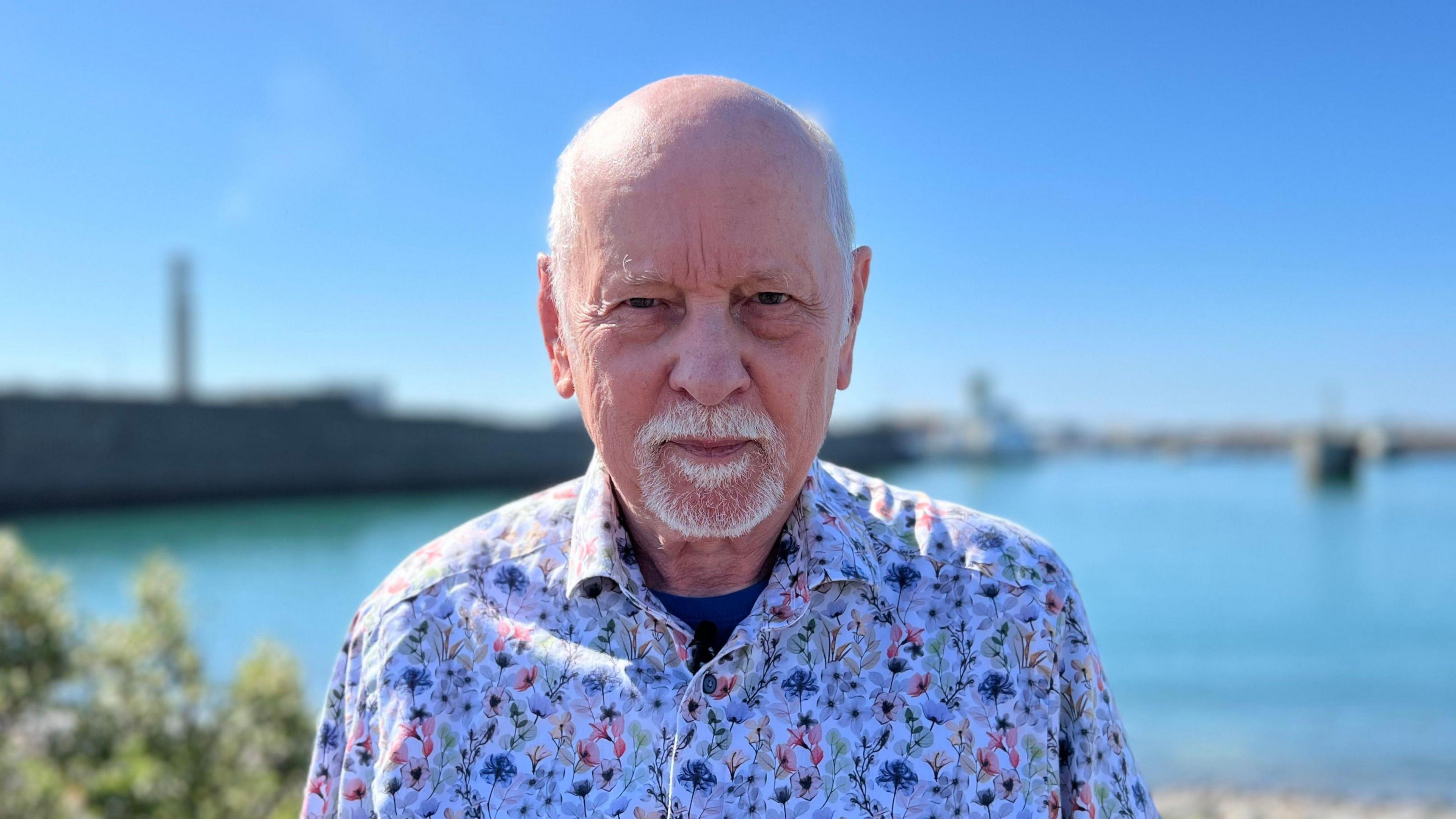 John Baker is staring into the camera while stood in front of Elizabeth Harbour. The sea and Albert Pier are out of focus behind him. He is wearing a white floral shirt.