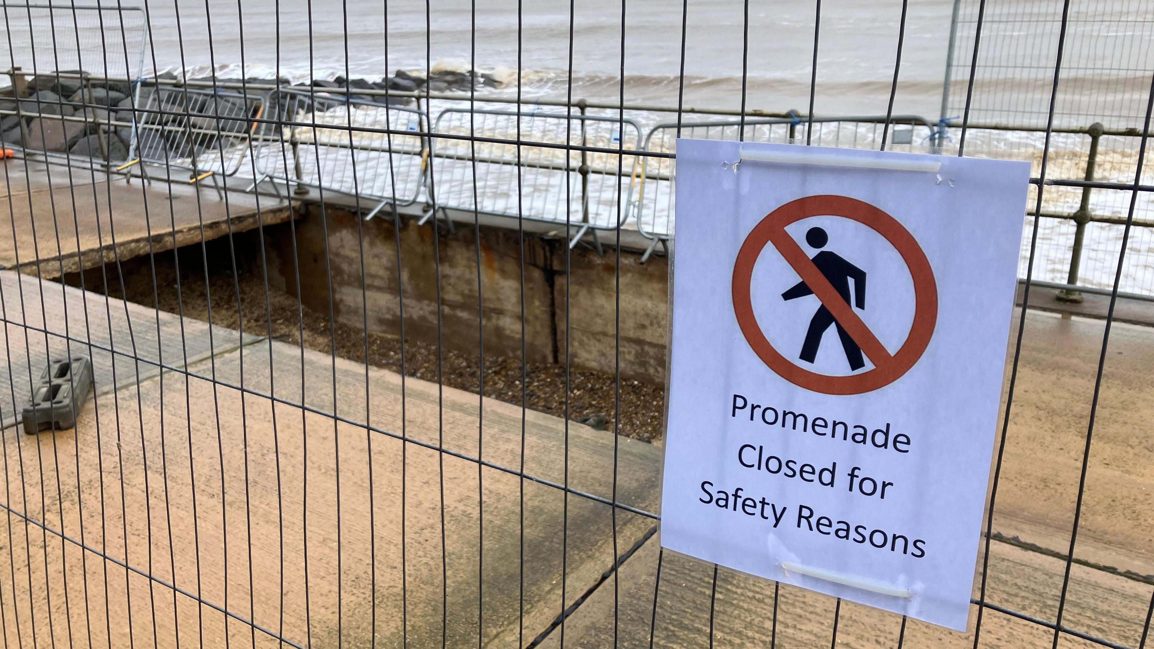 Metal fencing surrounds a large, rectangular-shaped hole in a concrete promenade with a sign on the fence reading "promenade closed for safety reasons". Waves and a rock groyne can also be seen in the background.