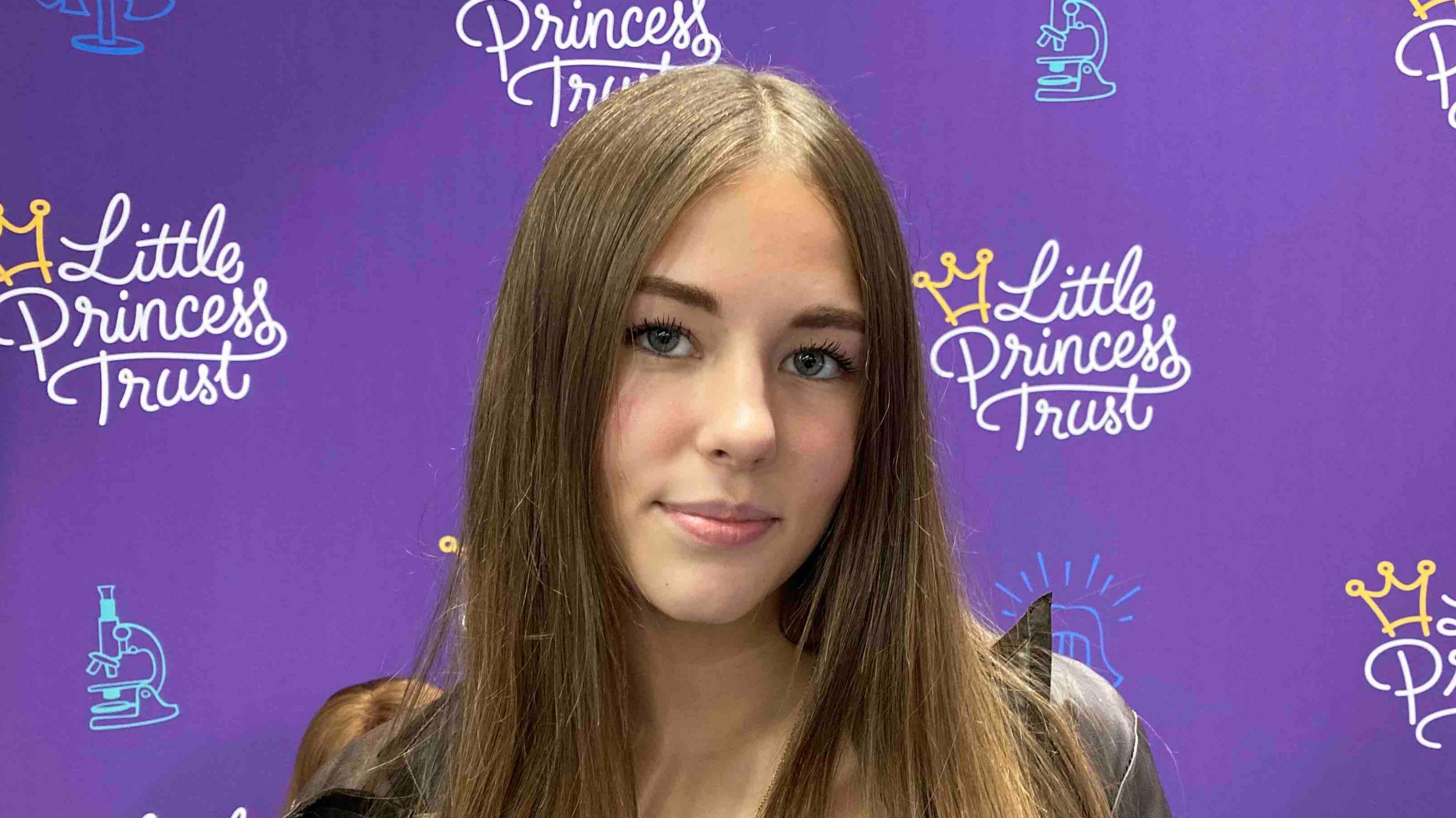 A girl with long brown hair is pictured in front of a purple background with the words Little Princess Trust written across it.