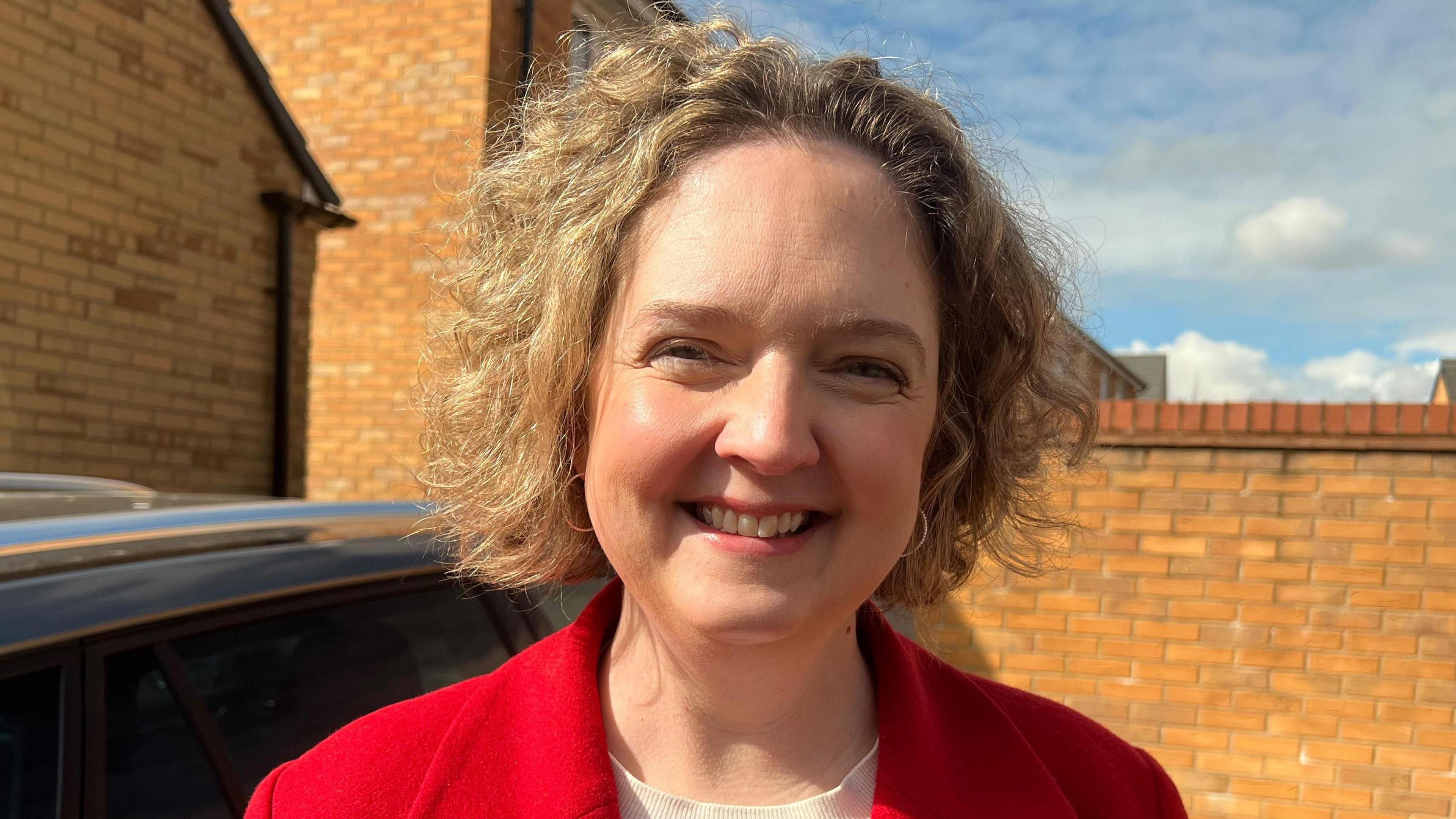 The picture shows Anna Sabine smiling into the camera. She is wearing a red jacket. There is a brick wall behind her and a car to the left of the picture.