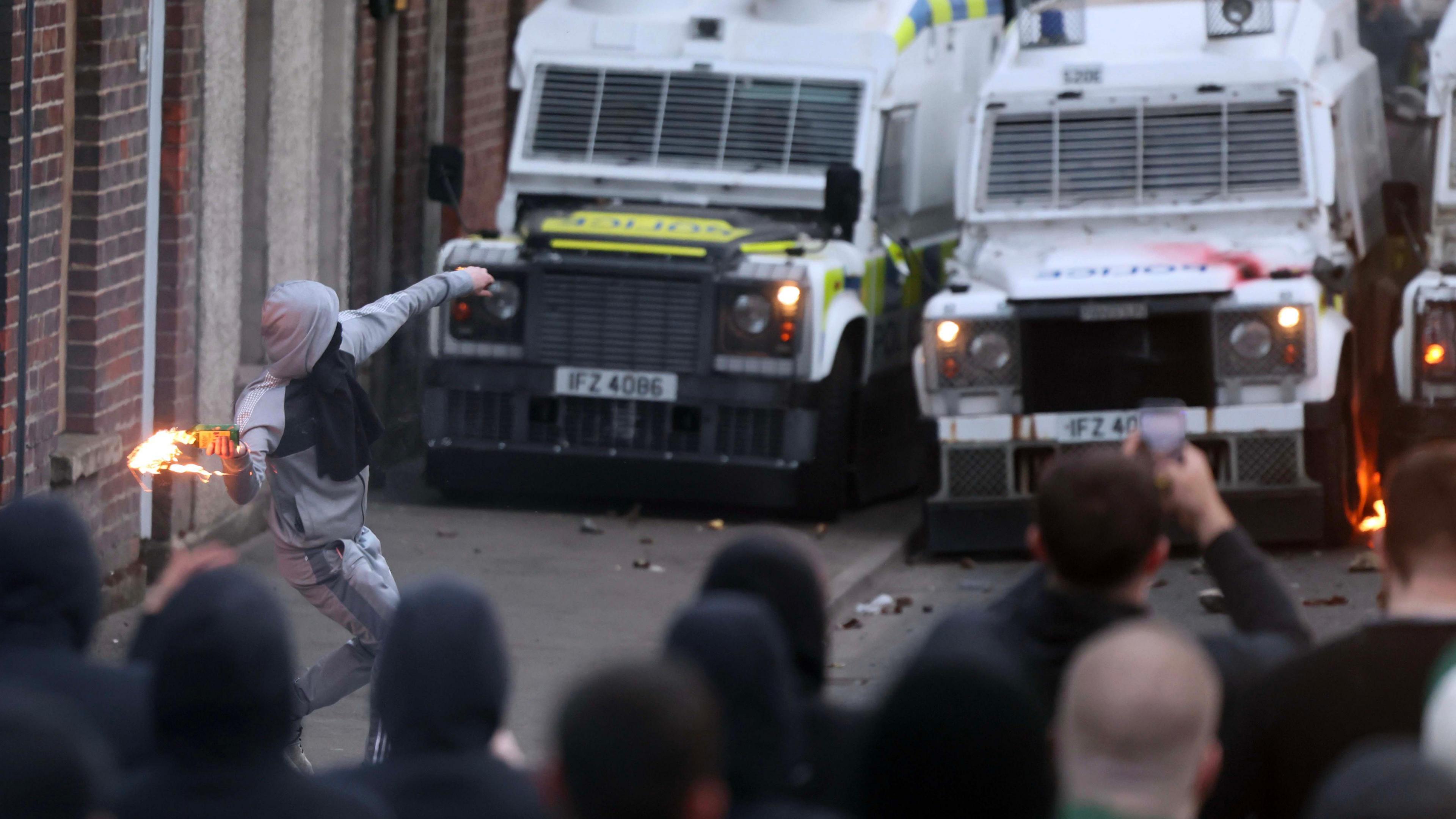 A make rioting is throwing a petrol bomb at a line of PSNI jeeps. A large number of people are facing the police cars. Debris is lying about the street.