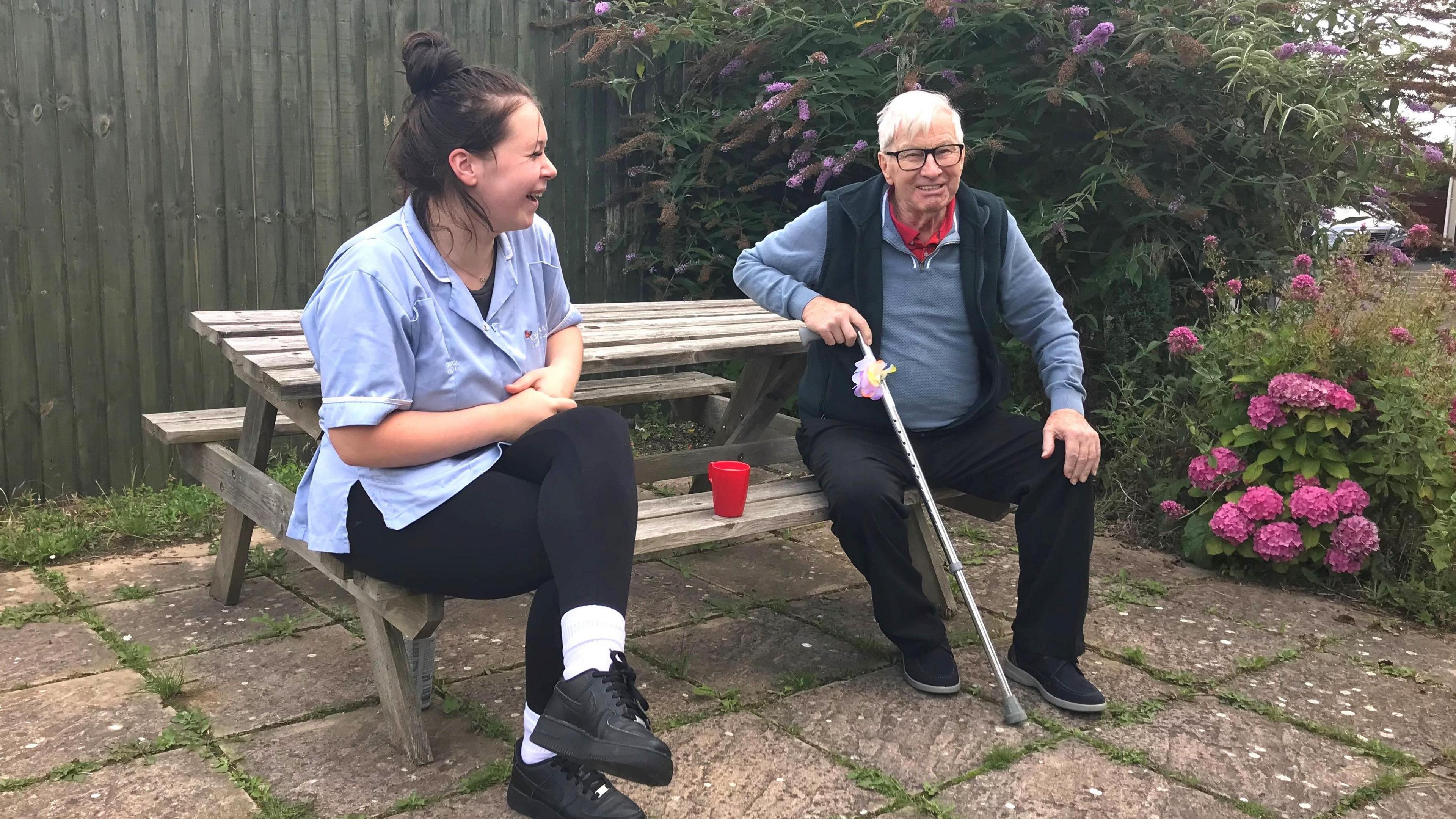 Chestnut Lodge resident Ted and carer Madi sit at a bench enjoying a cup of tea in the garden. 