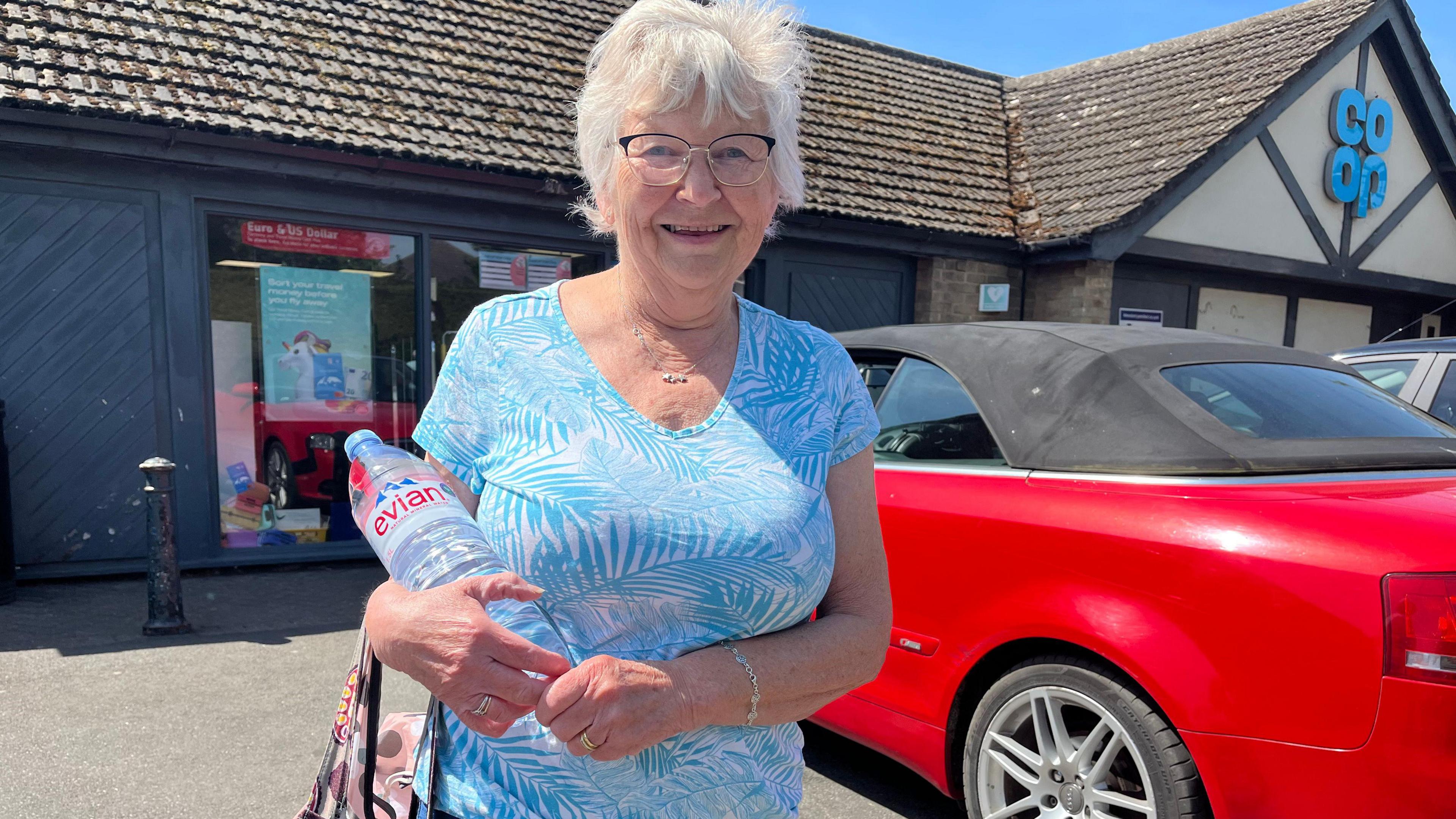 Margaret Smillie has shorty white hair and wears glasses. She is wearing a patterned white and blue top. She is holding a large bottle of evian water in her arms.