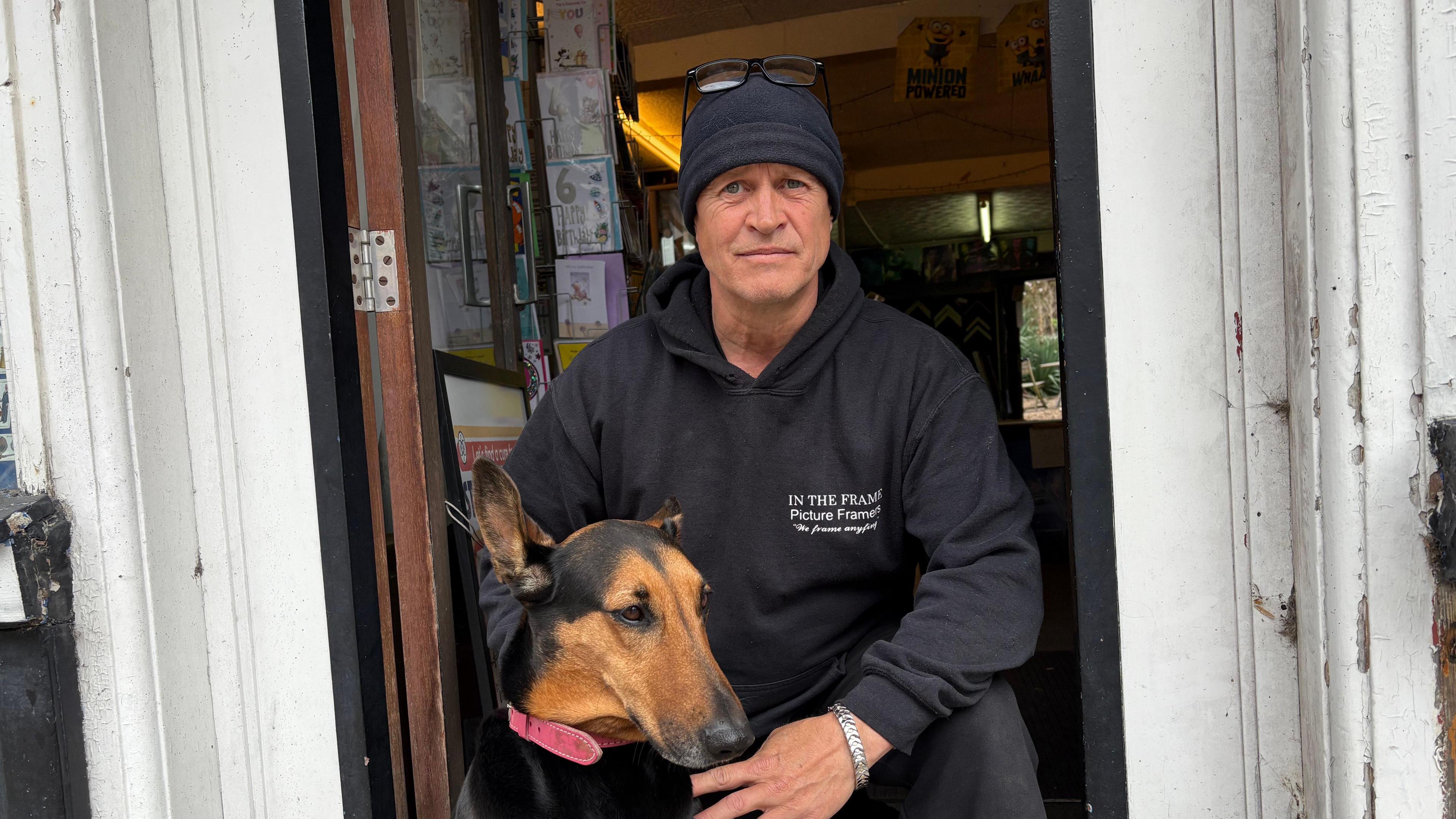 A man in a black hoodie, crouches in a doorway petting his dog.
