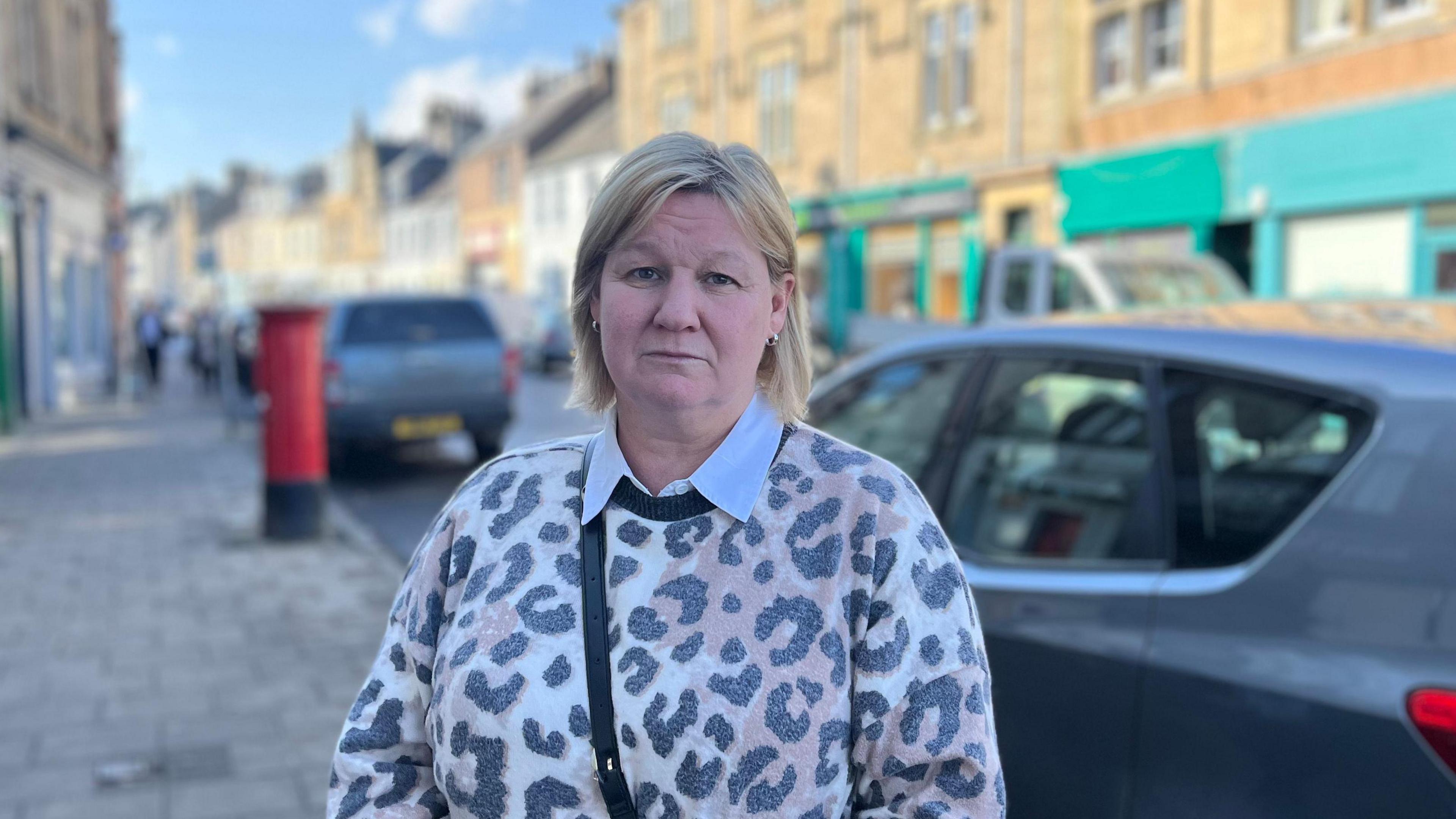 A blonde haired woman with animal-print top faces the camera with a street scene behind her