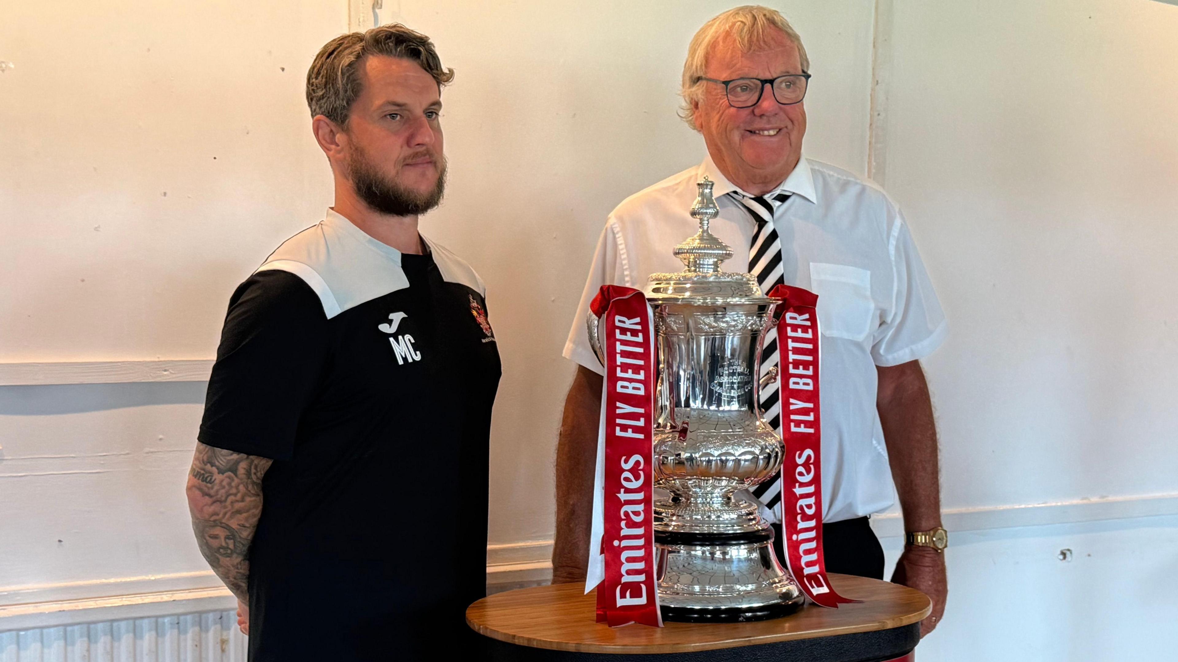 Two men, smiling, standing either side of the silver FA cup which has a red ribbon saying "Emirates fly better" tied to each handle. The man on the right is older and wearing a team tie that has black and white diagonal stripes. The man on the left is wearing a sports top with a red and yellow team logo on the chest.
