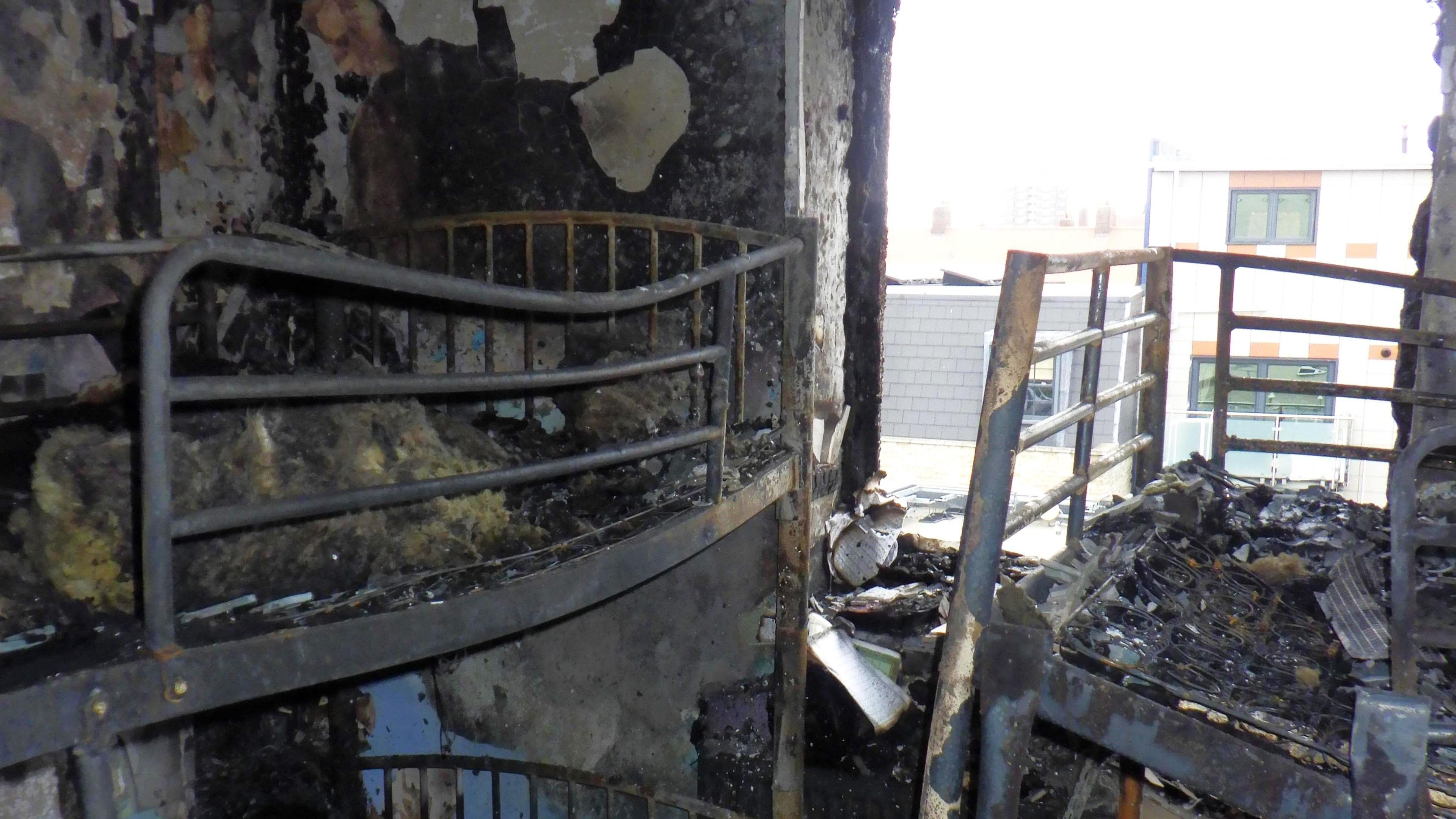 A view from inside the burned-out flat, with bunk bend apparently warped and twisted by the heat, with ash and debris where the mattresses would be and soot-blackened walls. Where the window would have been, the flat is now open to the elements.