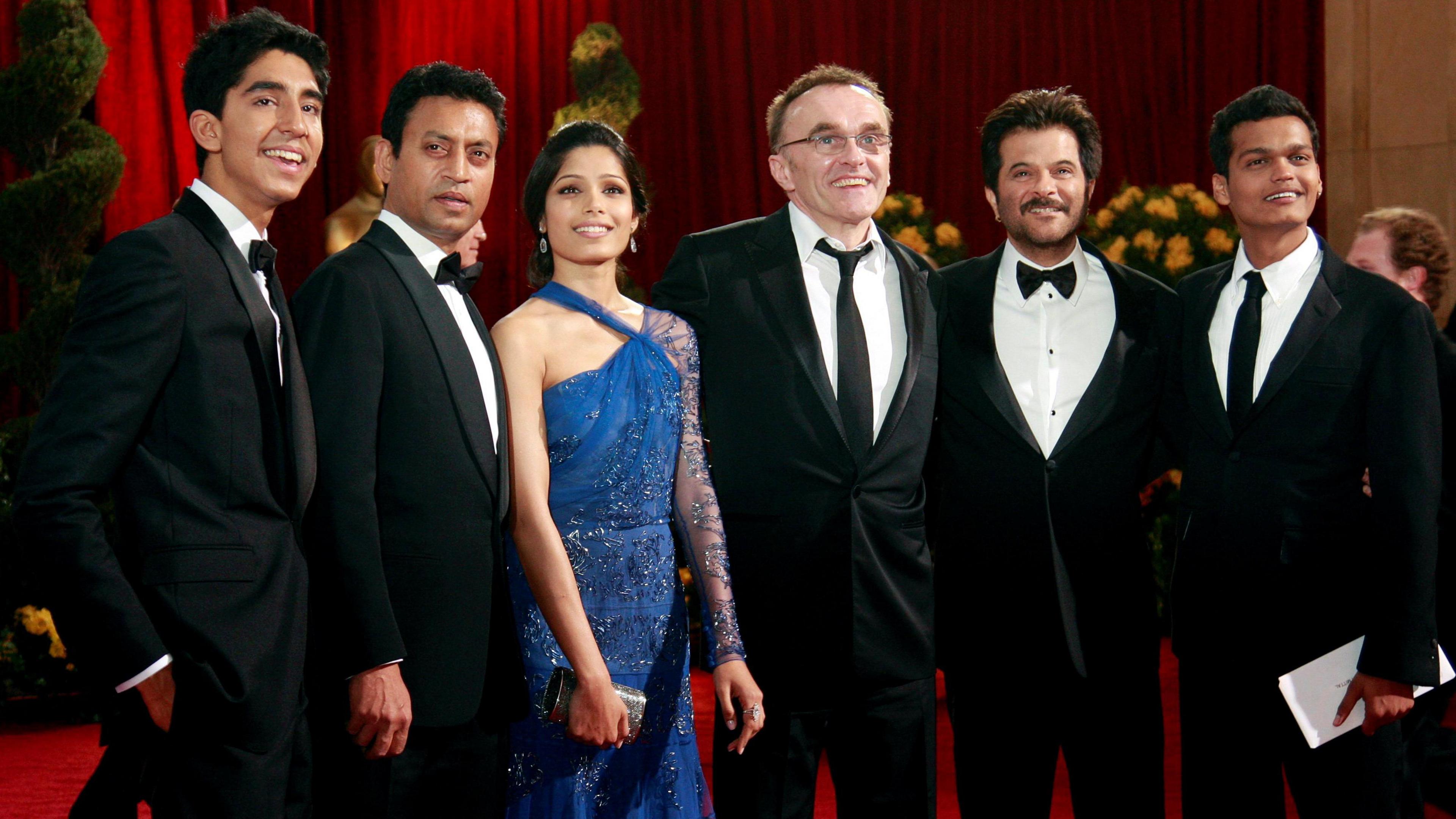 Slumdog Millionaire cast members gather at the Oscars. Dev Patel, Irrfan Khan, Freida Pinto on the left of Danny Boyle, with Anil Kapoor and Madhur Mittal on his right. They pose on the red carpet before the Oscars ceremony. The men are wearing smart black suits and ties, while Freida Pinto is in a blue sparkly dress.
