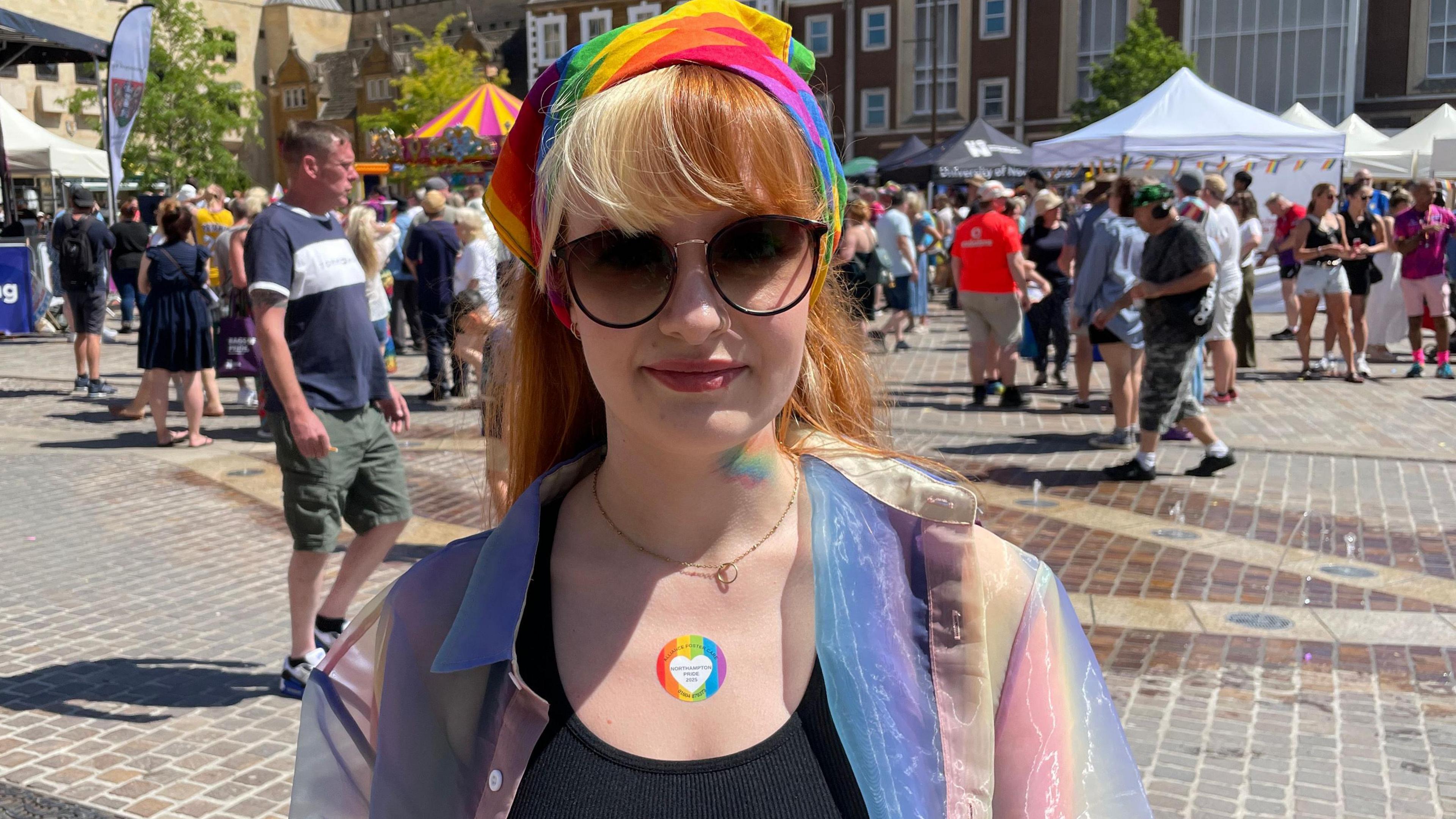 Kamila is standing in the Market Square in front of gazebos and crowds of people. She is wearing sunglasses, a rainbow headscarf, black top and purple, green and yellow type of mesh jacket. She is smiling at the camera. She has long hair and a fringe. Her hair is blonde on one side and orange/ ginger on the other.