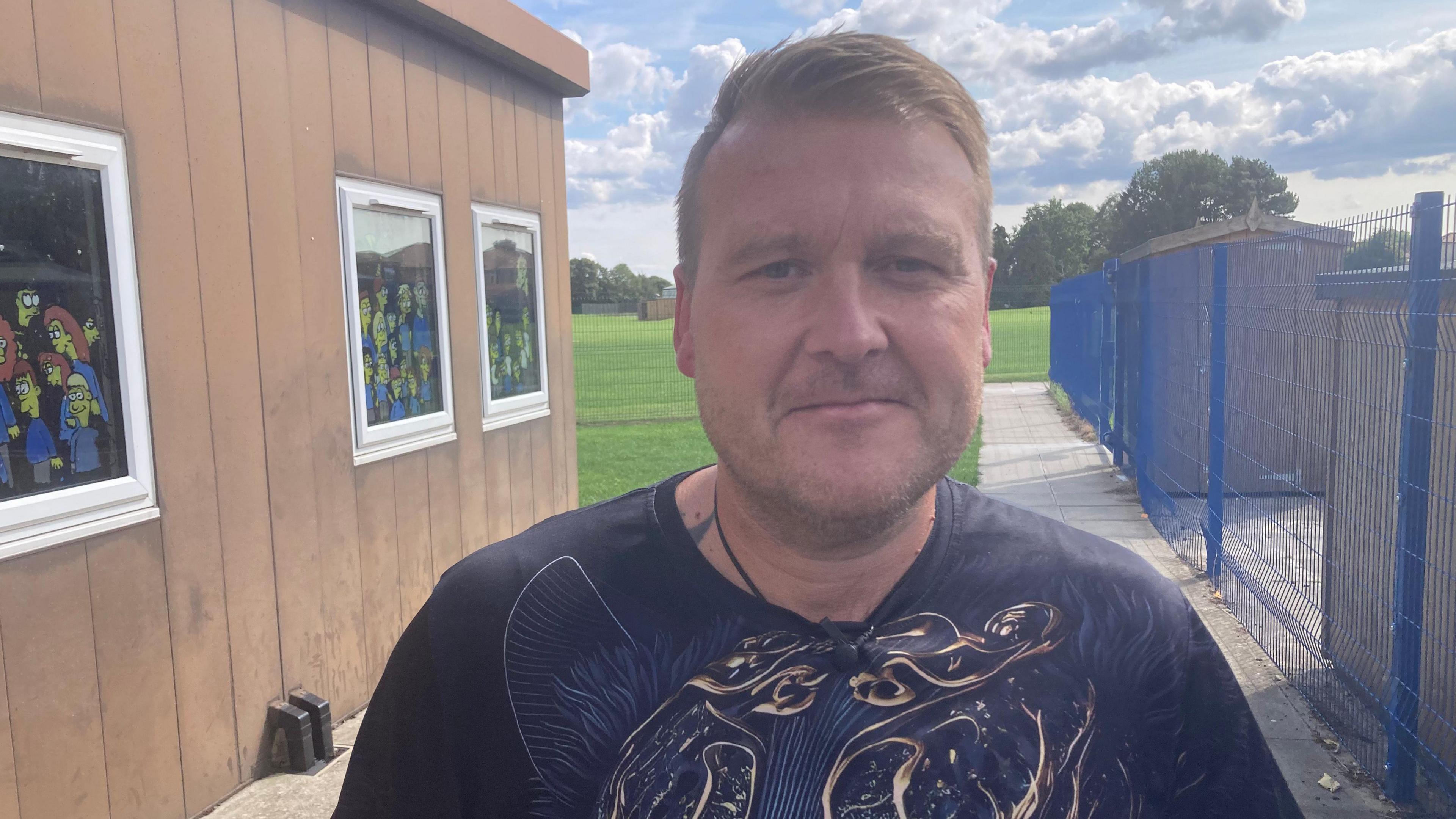 A man in a black t-shirt stands next to a school building.