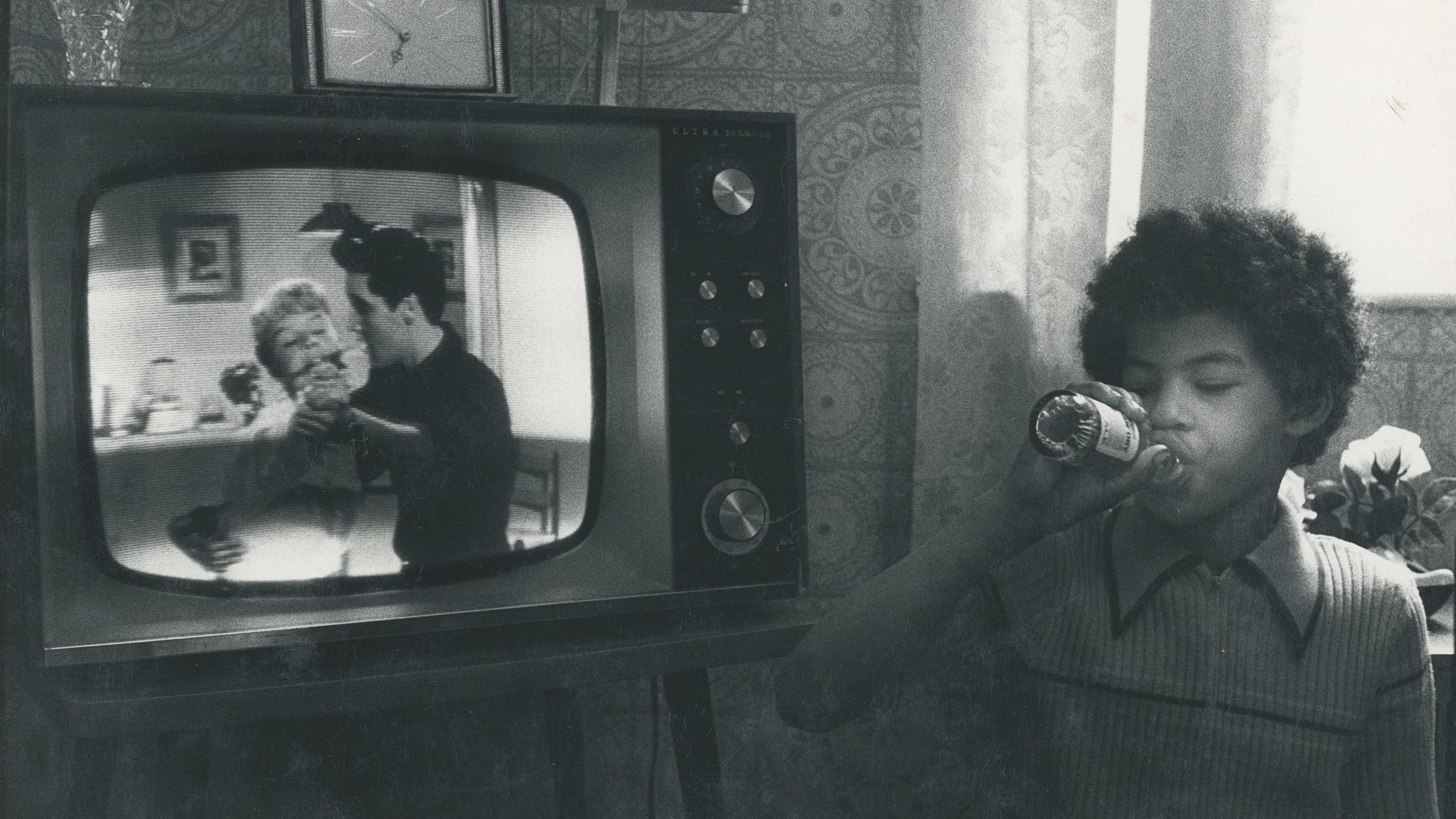 A black and white image of a boy sitting in front of a television drinking from a soda bottle.