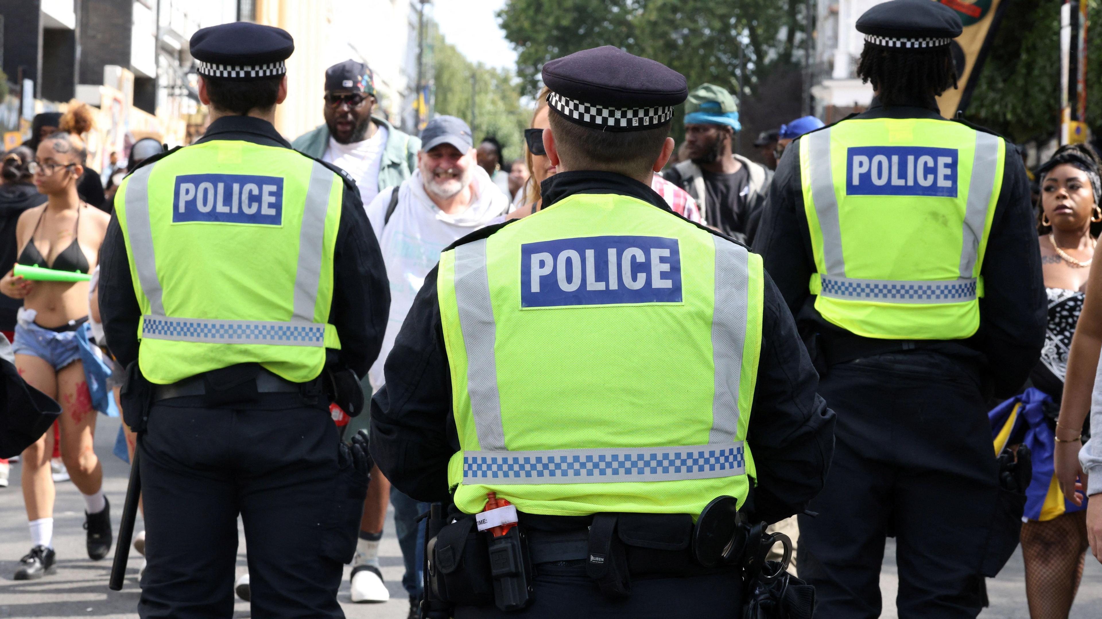 The backs of three police officers in green high viz uniform face carnival-goers walking towards them