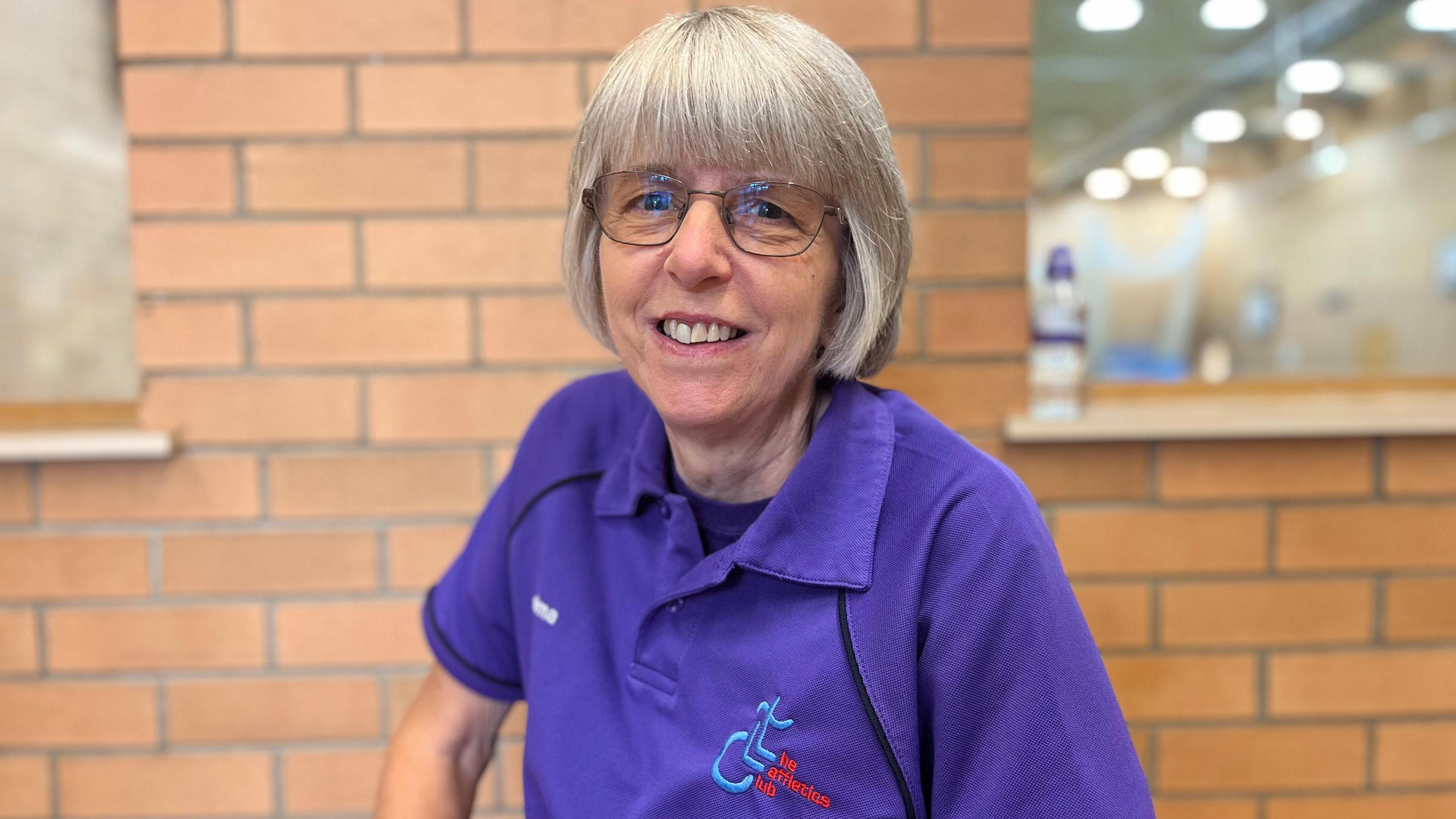 Thelma Smith is smiling at the camera. She is wearing a purple T-shirt with 'The Laffletics Club' printed on it. She has a short grey bob and is wearing glasses. She is sitting in front of a brick wall. 