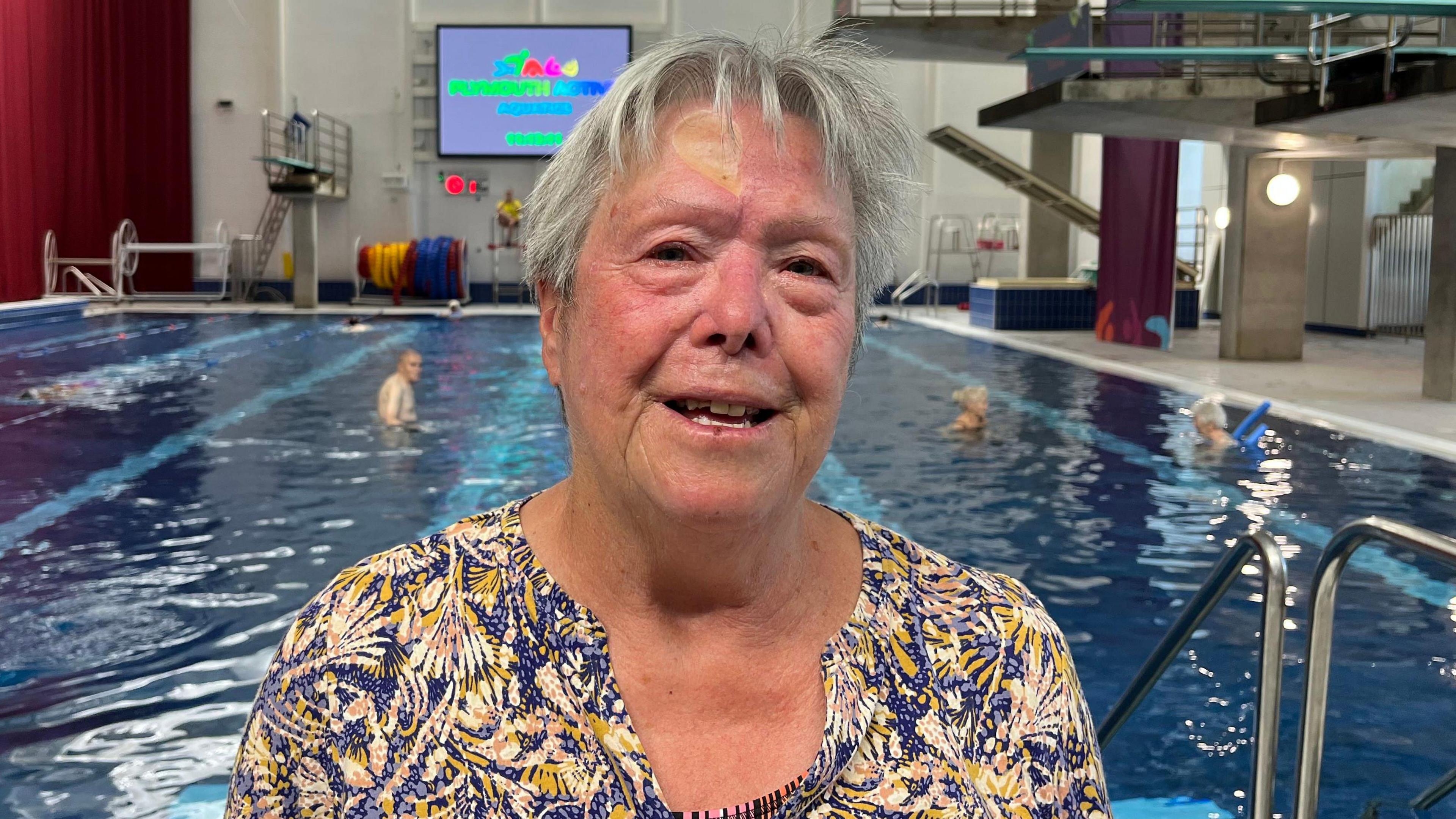 A woman with grey hair and a shirt with a yellow and blue pattern. In the background is a swimming pool.